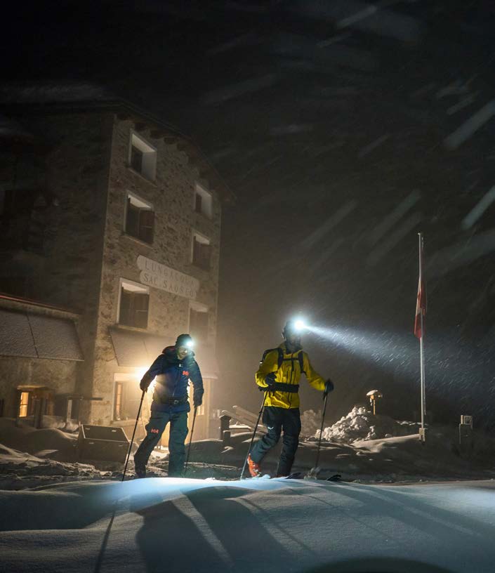 Zwei Skitourengeher mit Stirnlampen vor einer Berghütte in der Dämmerung