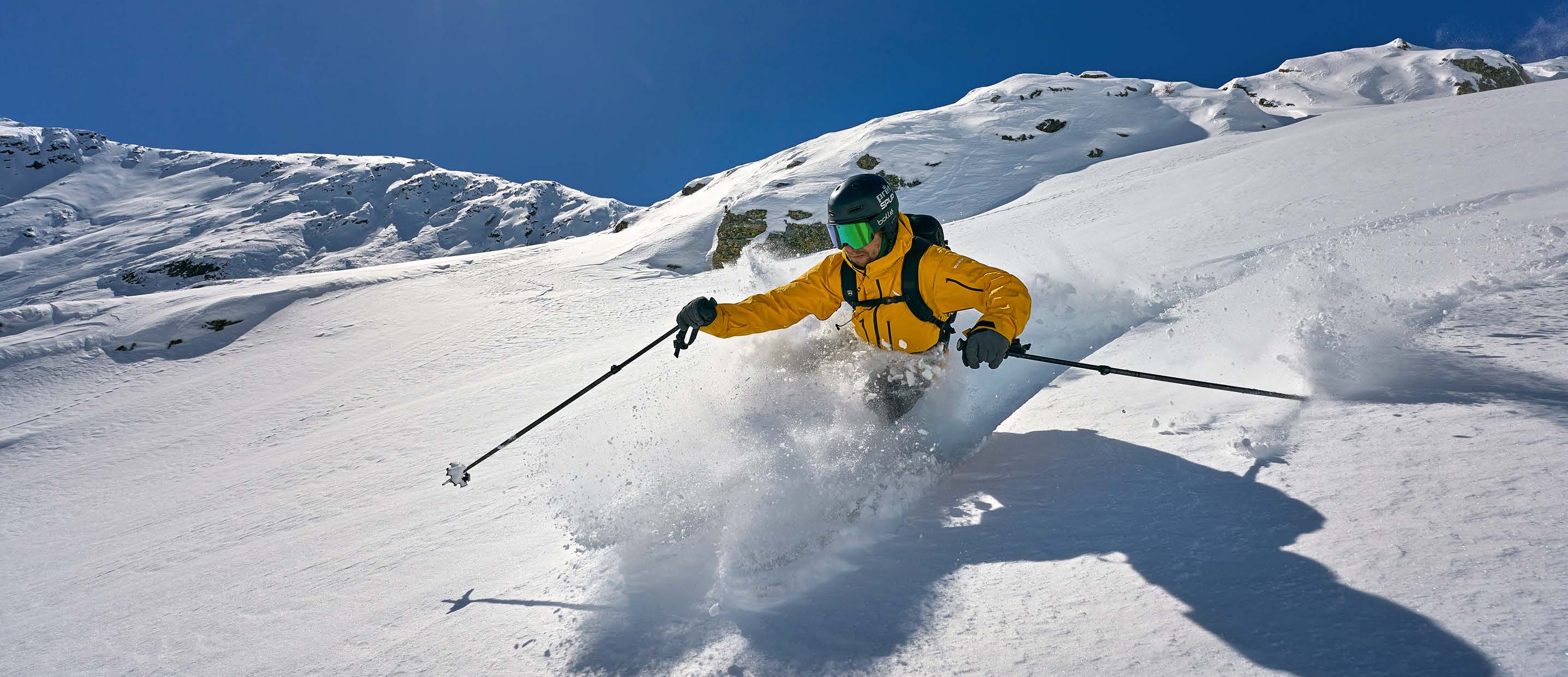 Skifahrer in gelber Jacke zieht einen Pulverschneeschwung in sonnigem Gelände