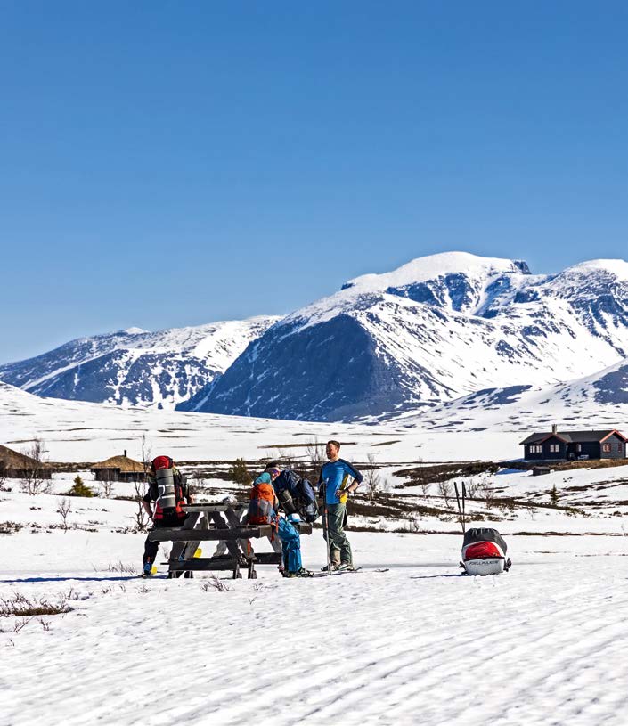 Drei Skitourengeher an einem Picknicktisch vor verschneiten Rondane-Gipfeln