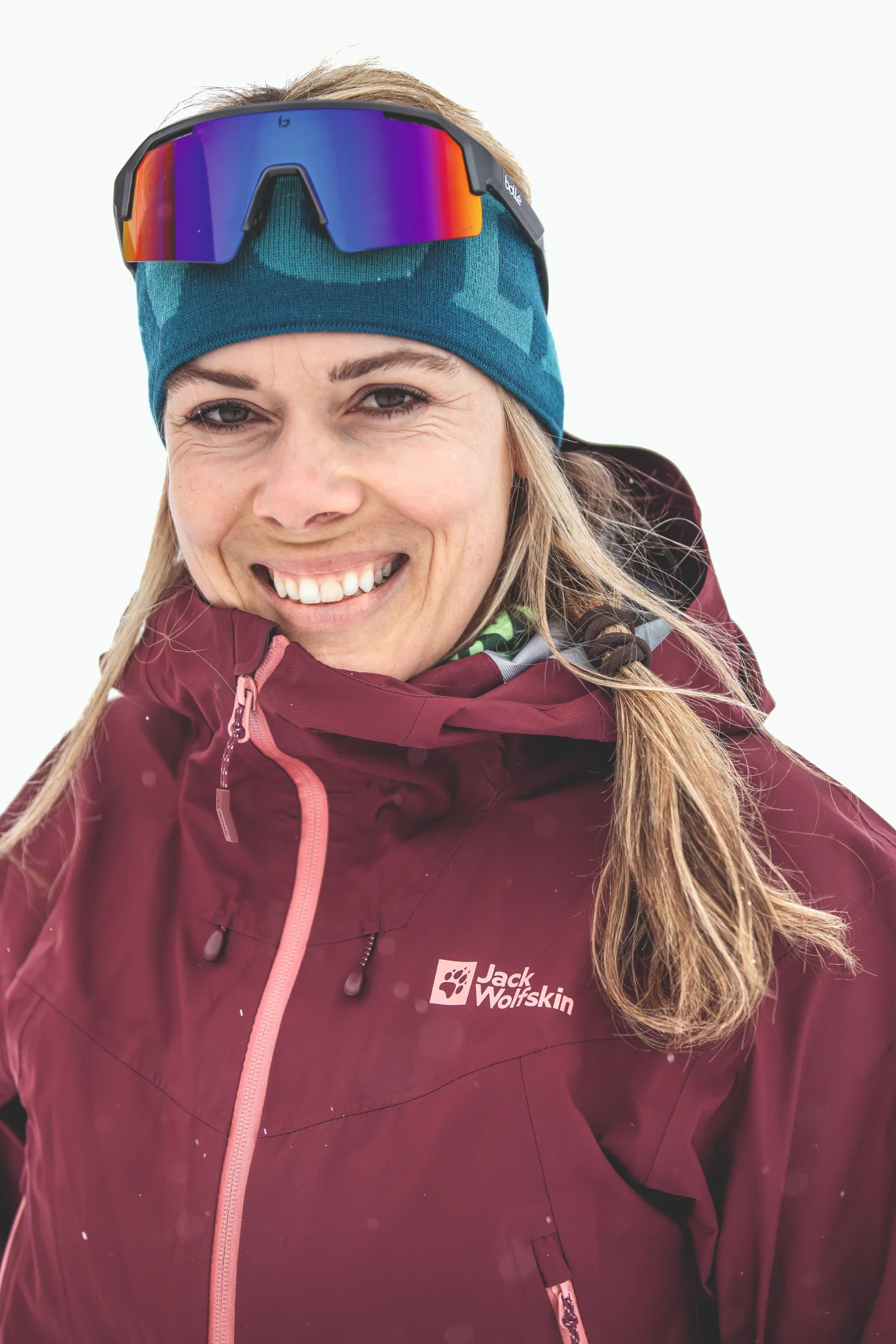 Andrea Limbacher lächelt in Nahaufnahme vor weißem Hintergrund, mit petrolfarbenem Stirnband, darauf sitzender verspiegelter Sportbrille und burgunderroter Jacke.
