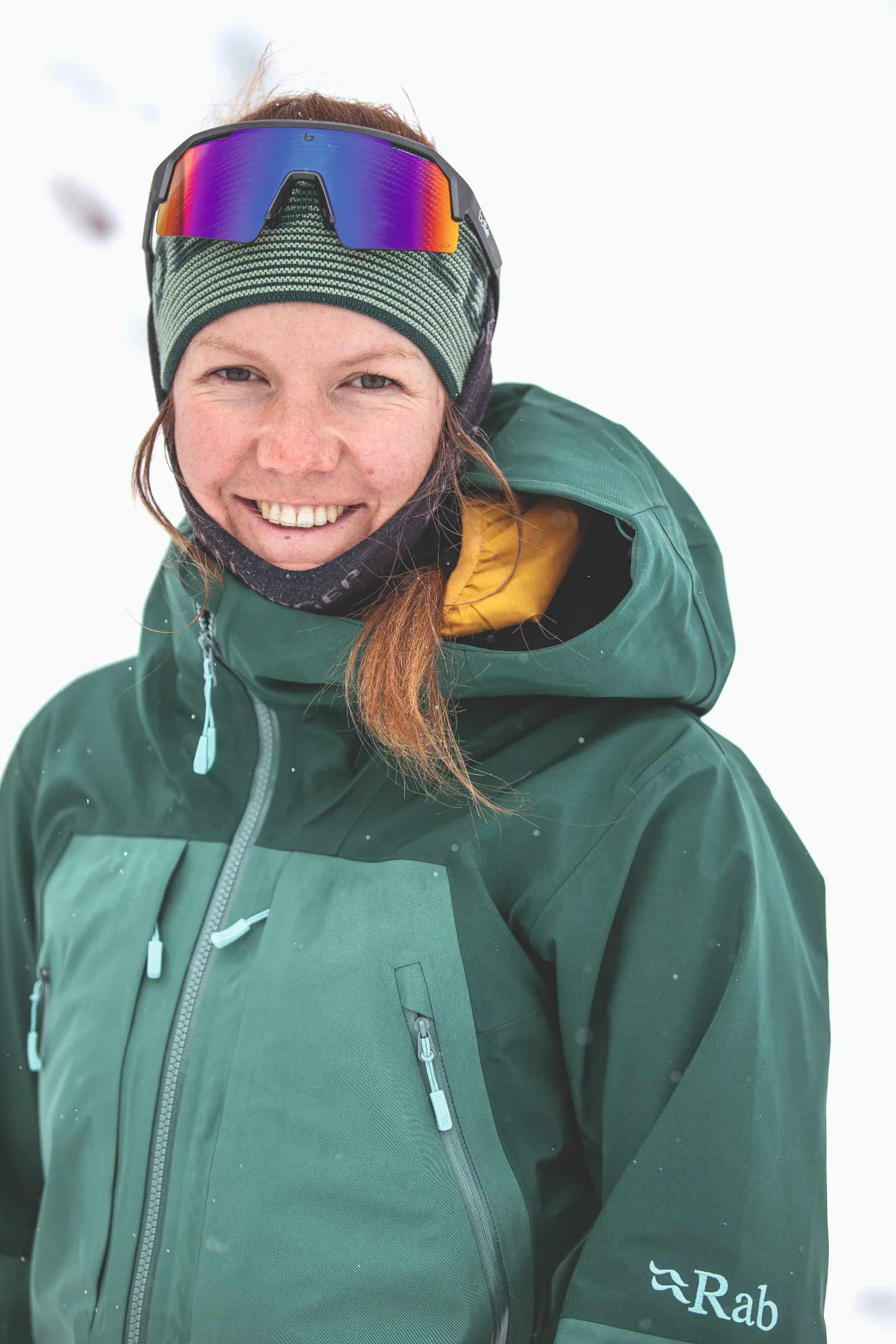 Annika Schlachter lächelt vor verschneitem Hintergrund in die Kamera, mit grüner Jacke, gestreiftem Stirnband und einer verspiegelten Sportbrille auf der Stirn.