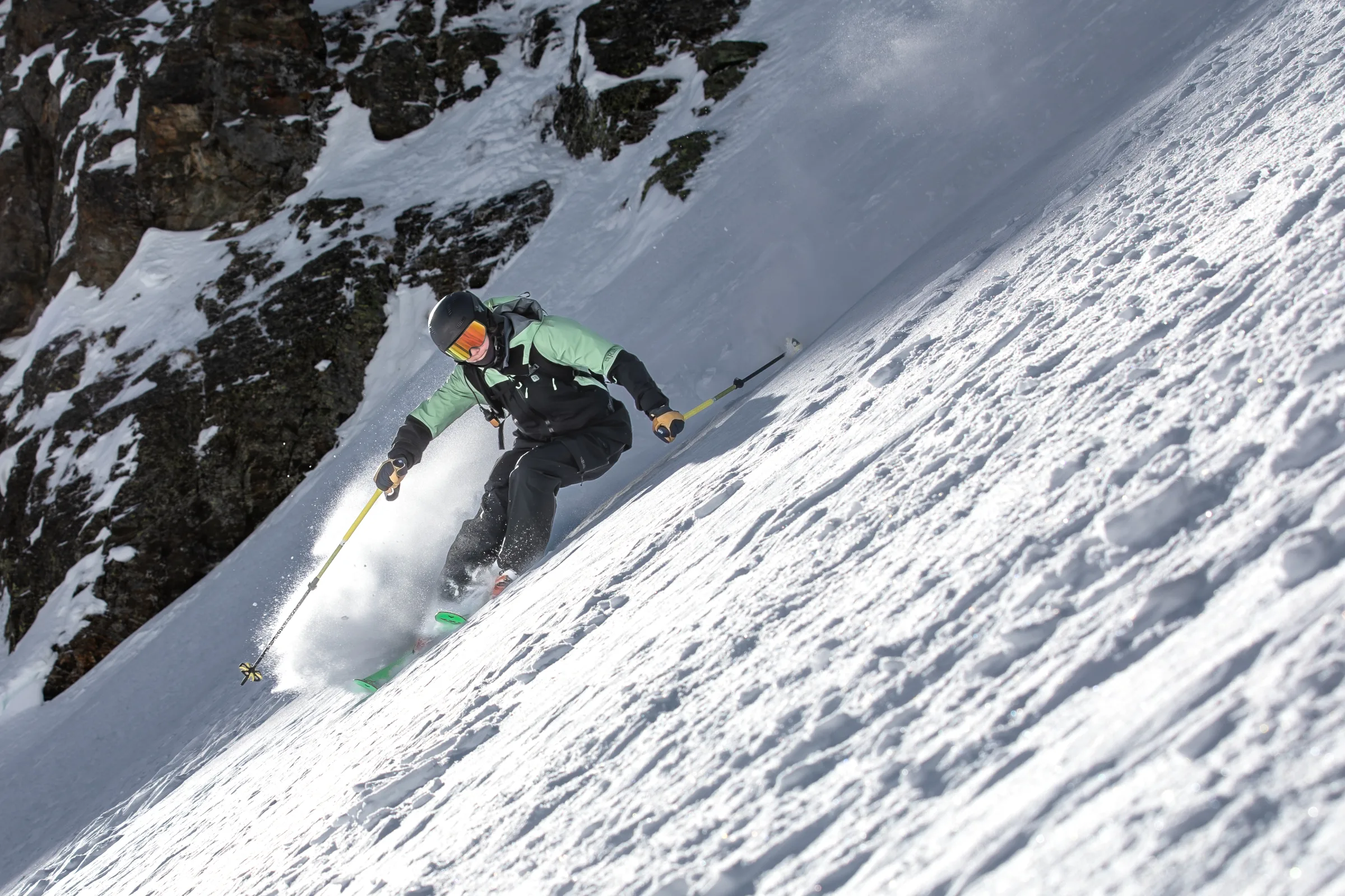 Annika Schlachter fährt mit grünen Ski und gelben Stöcken einen steilen Schneehang neben dunklen Felswänden hinab und wirbelt Schnee auf.