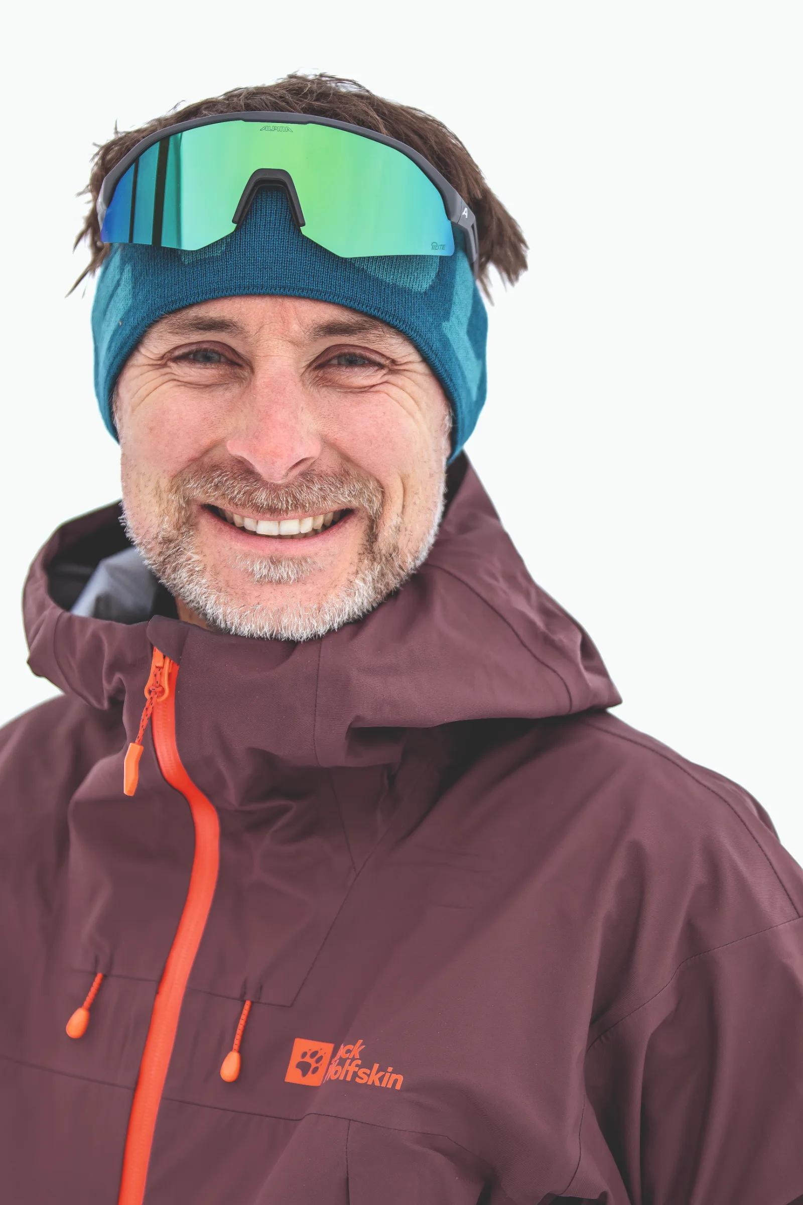 Bernd Hassmann lächelt vor weißem Hintergrund in die Kamera, mit blauem Stirnband, hochgeschobener grün verspiegelter Sportbrille und weinroter Jacke.