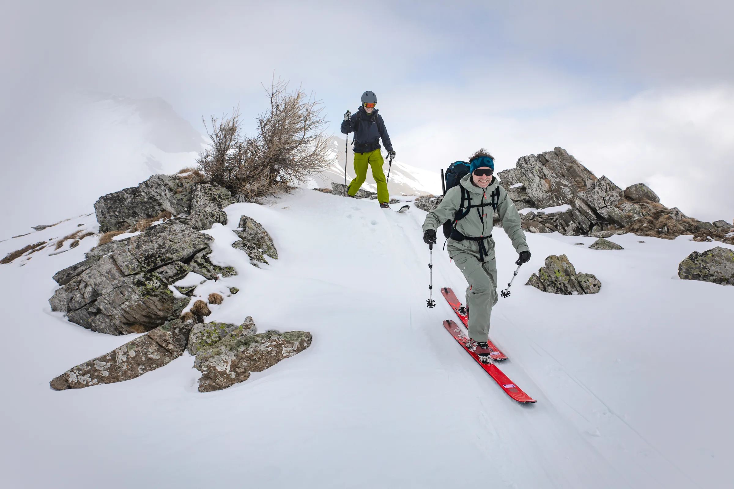 Bernd Hassmann geht auf roten Ski zwischen Felsen über einen verschneiten Grat bei Lenzerheide, gefolgt von einer weiteren Person im Nebel.