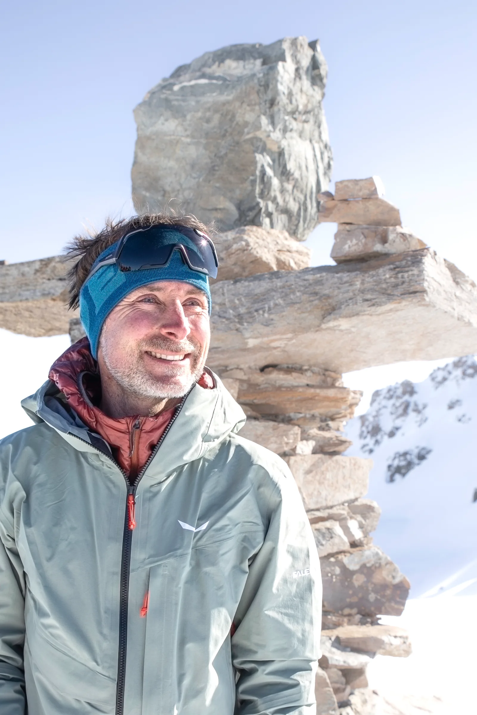 Bernd Hassmann lächelt mit blauem Stirnband und hochgeschobener Sonnenbrille vor aufgeschichteten Steinplatten und schneebedeckten Bergen.