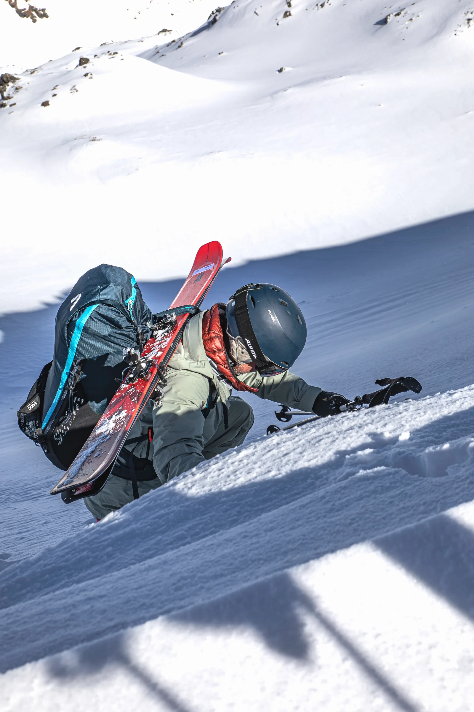 Bernd Hassmann steigt mit Helm und roten Ski am Rucksack einen steilen Schneehang hinauf und stützt sich mit einer Hand im Schnee ab.