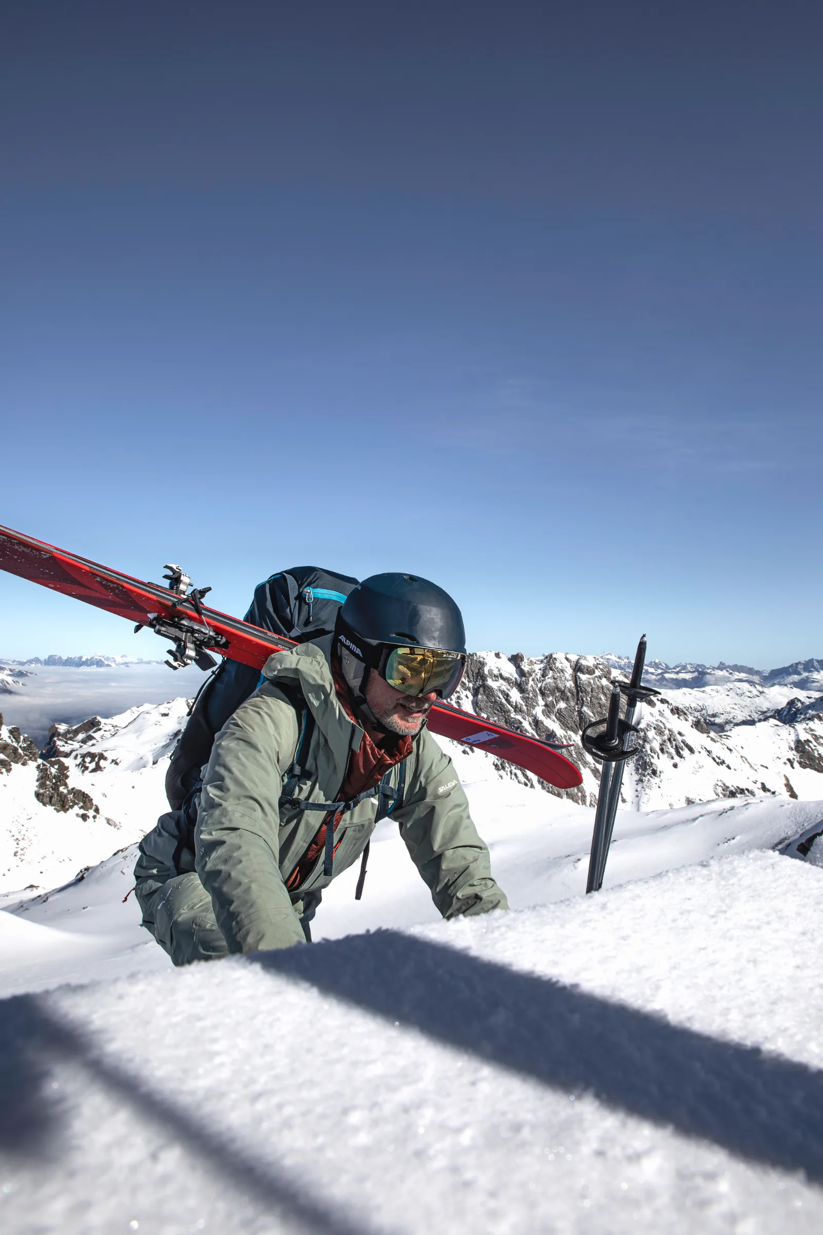 Bernd Hassmann klettert mit roten Ski quer am Rucksack über eine Schneekante; neben ihm stecken Stöcke, dahinter liegen verschneite Gipfel unter blauem Himmel.