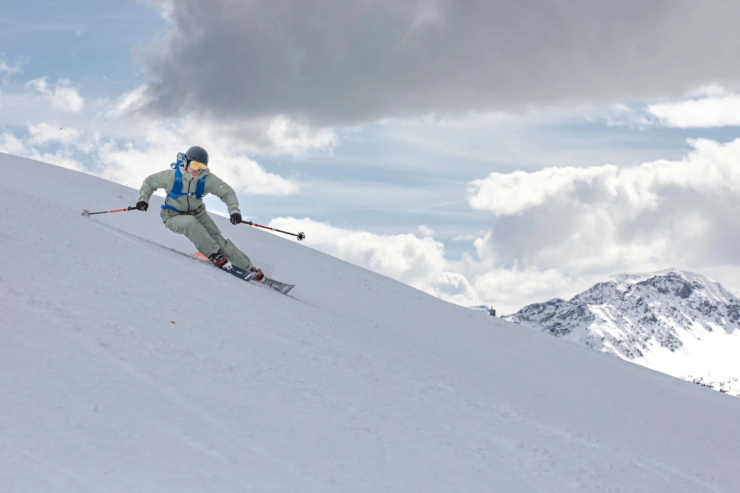 Bernd Hassmann legt sich mit blauem Rucksack in einen Schwung auf einem offenen Schneehang, während Schnee aufwirbelt und Wolken über einem Berggipfel stehen.