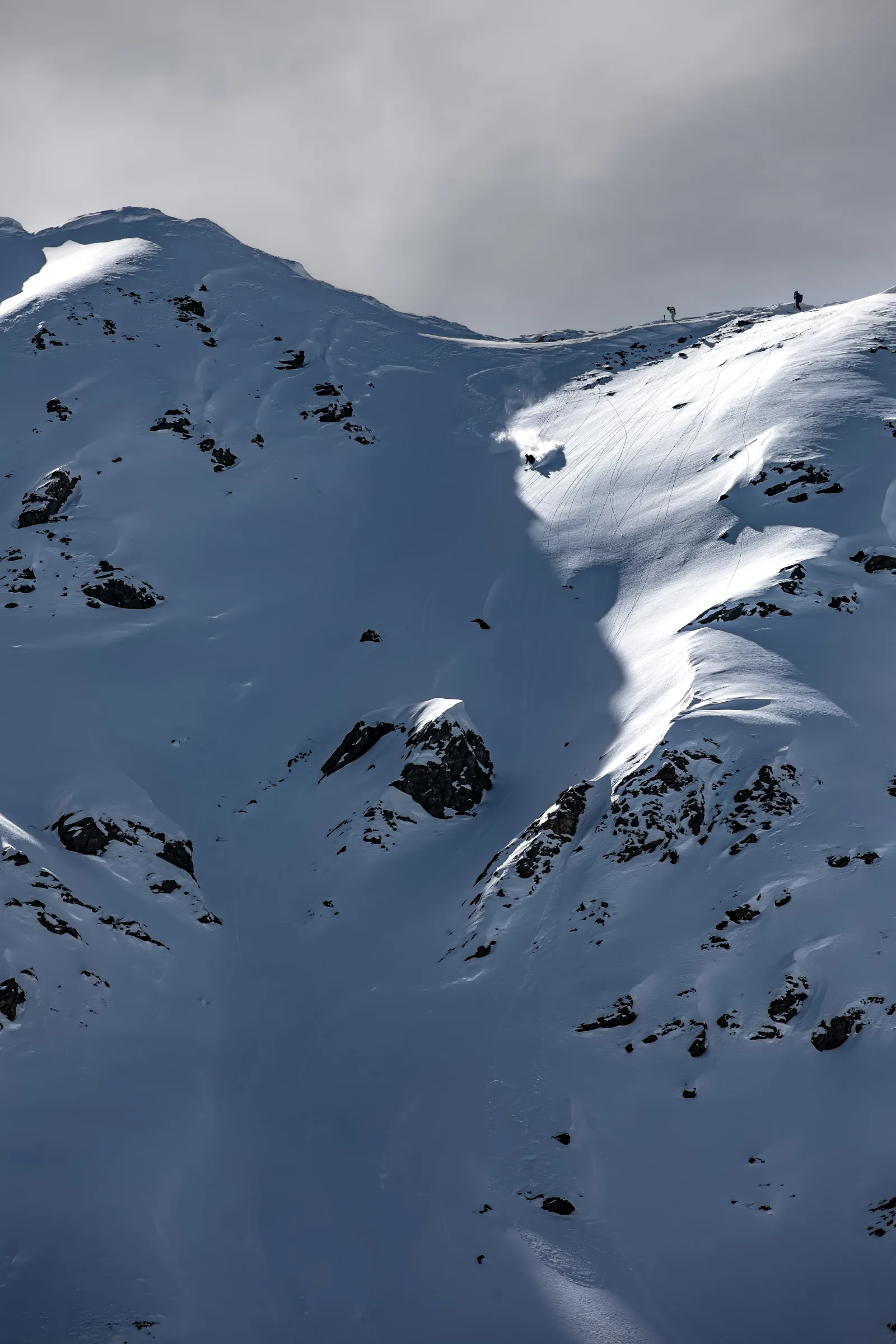 Ein Skifahrer zieht mit aufsprühendem Schnee einen Schwung durch einen steilen, felsdurchsetzten Hang, während zwei Personen oben am Grat stehen.