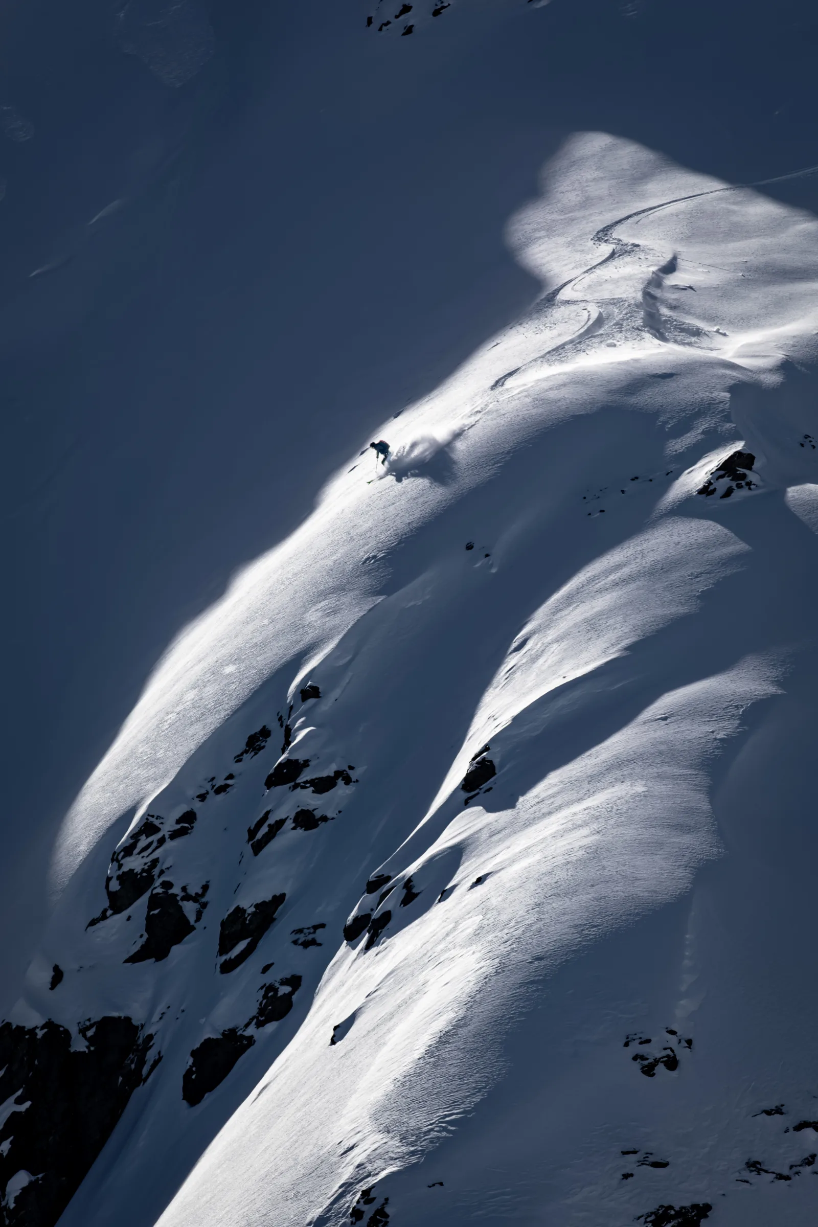 Ein Skifahrer fährt durch einen schmalen Sonnenstreifen am steilen Schneehang und hinterlässt eine geschwungene Spur zwischen Schatten und Felsen.