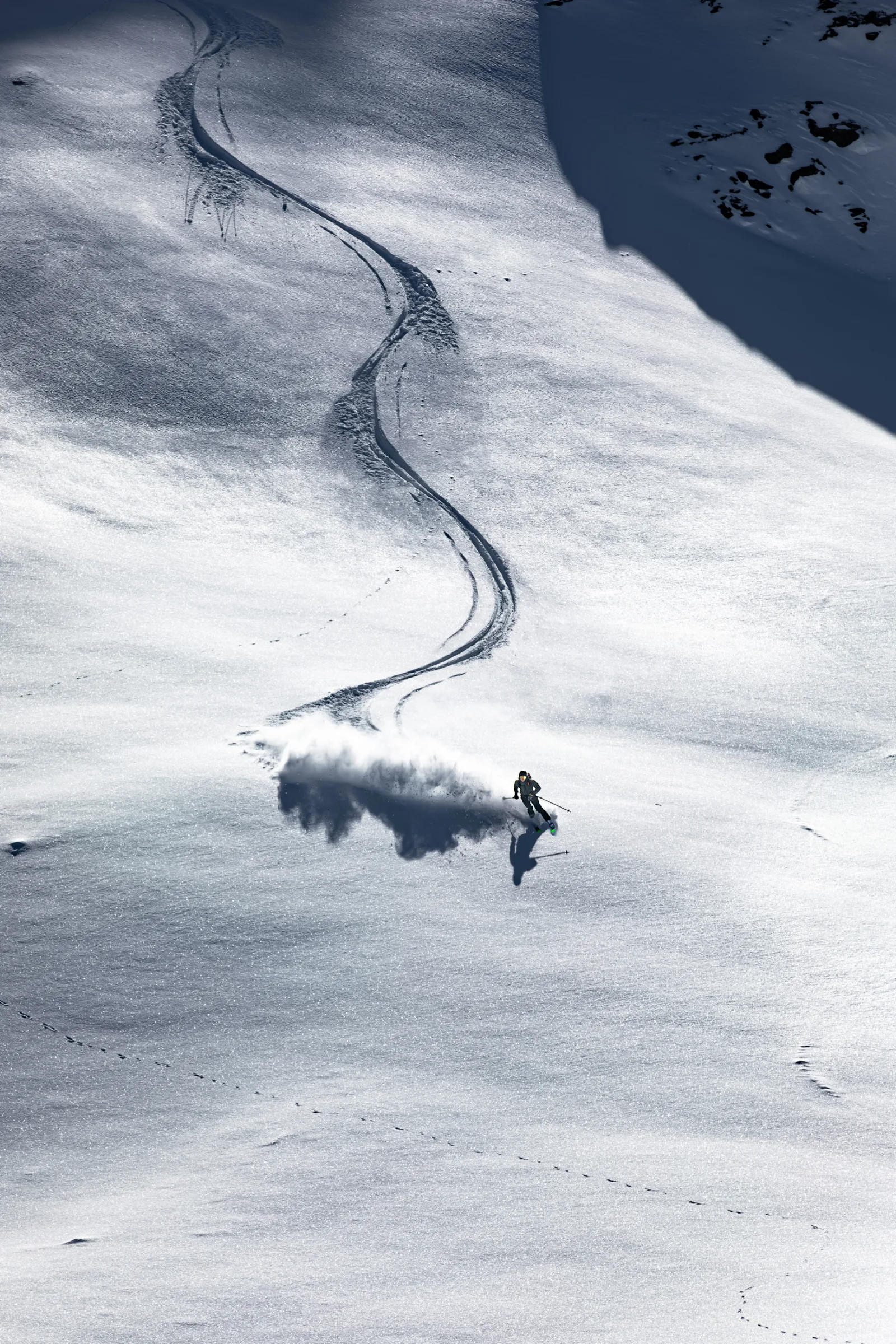 Ein Skifahrer wirbelt auf einem offenen, sonnigen Schneehang eine breite Schneewolke auf; eine lange S-förmige Spur zieht sich hinter ihm bergauf.