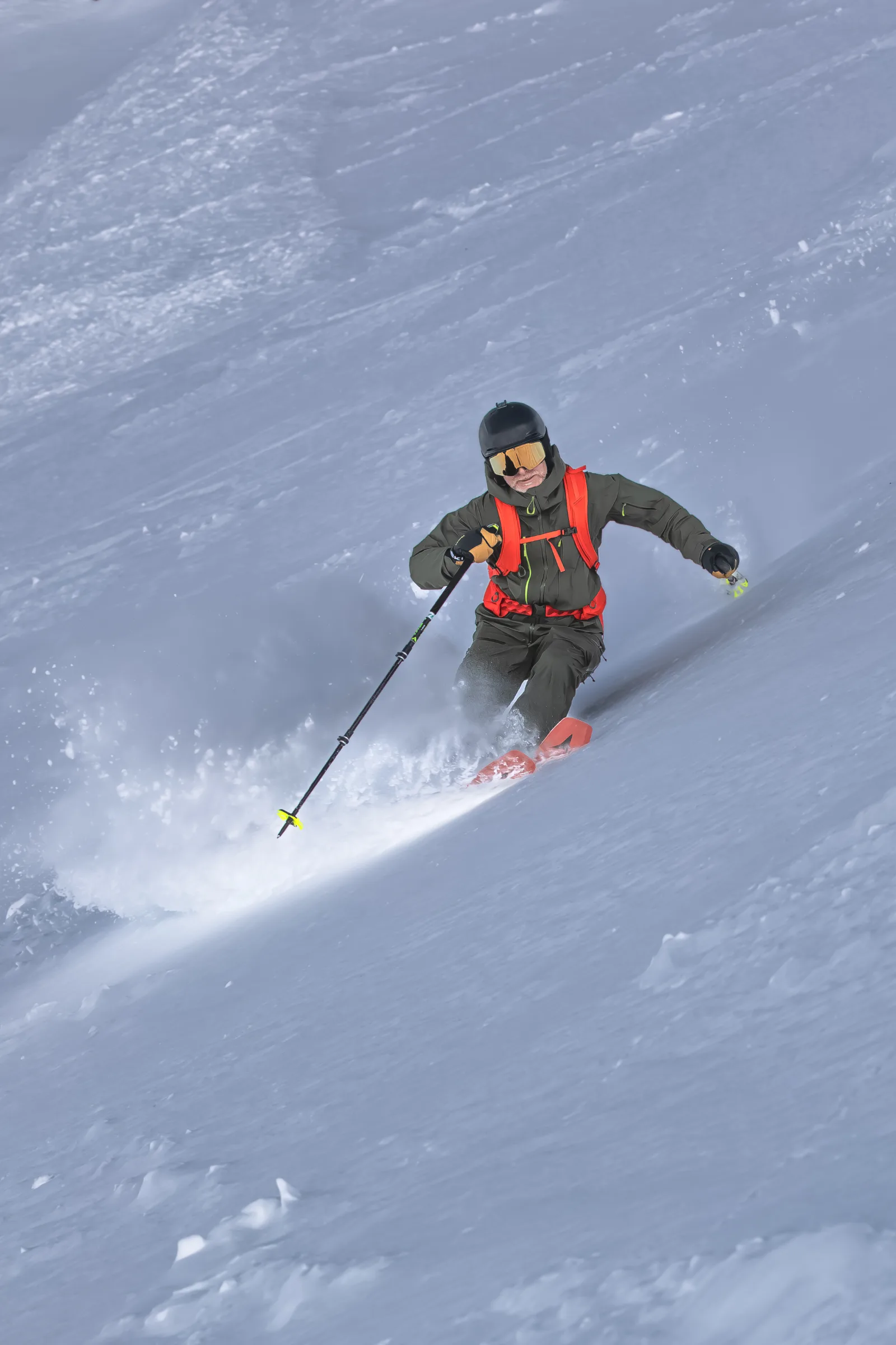 Bernd Hassmann fährt mit schwarzem Helm, verspiegelter Brille und orangefarbenem Rucksack durch tiefen Schnee, der um seine roten Skispitzen aufstiebt.
