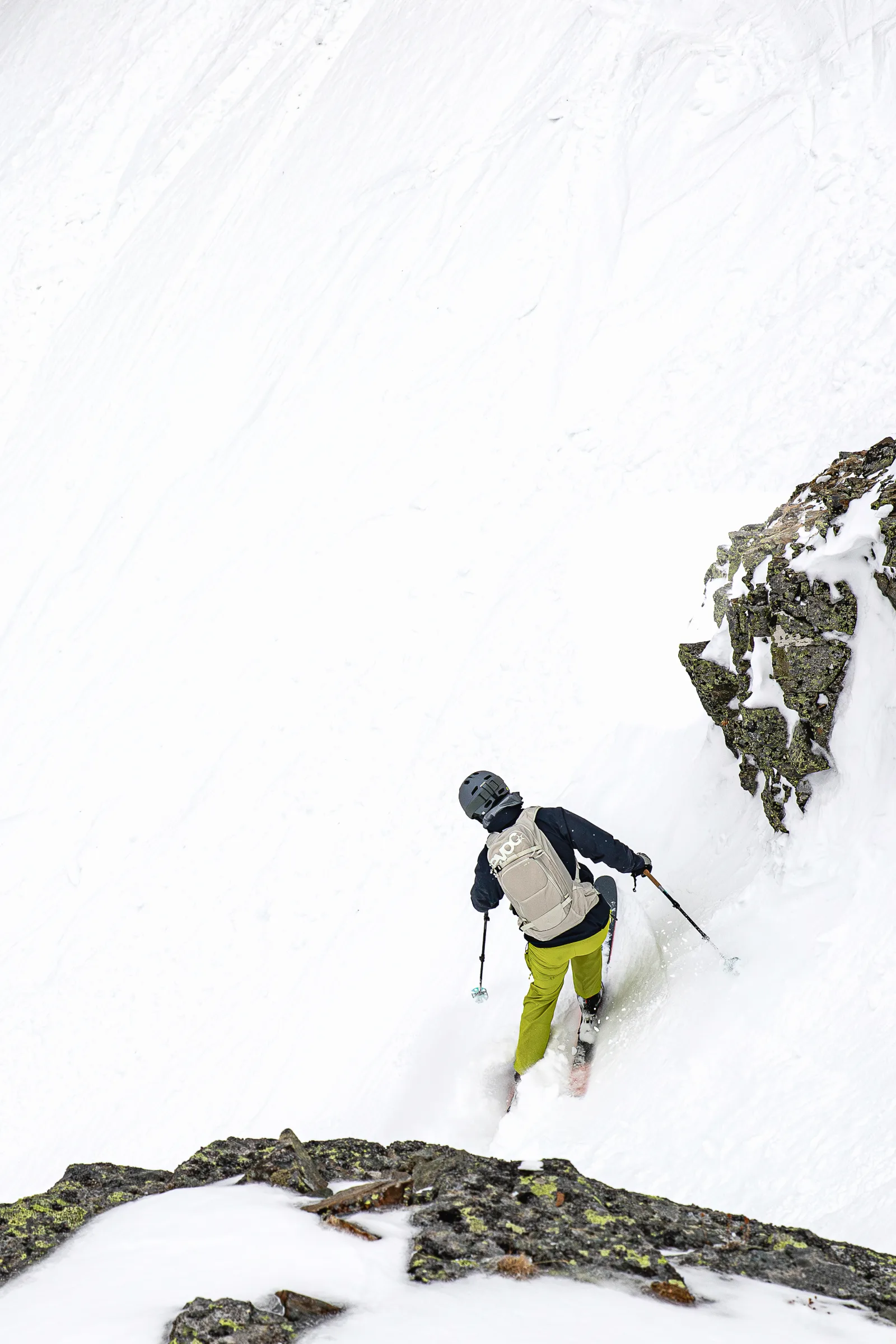 Von hinten gesehen fährt Christoph Öfele mit hellem Rucksack und gelbgrüner Hose einen steilen Schneehang neben einer Felswand hinab.