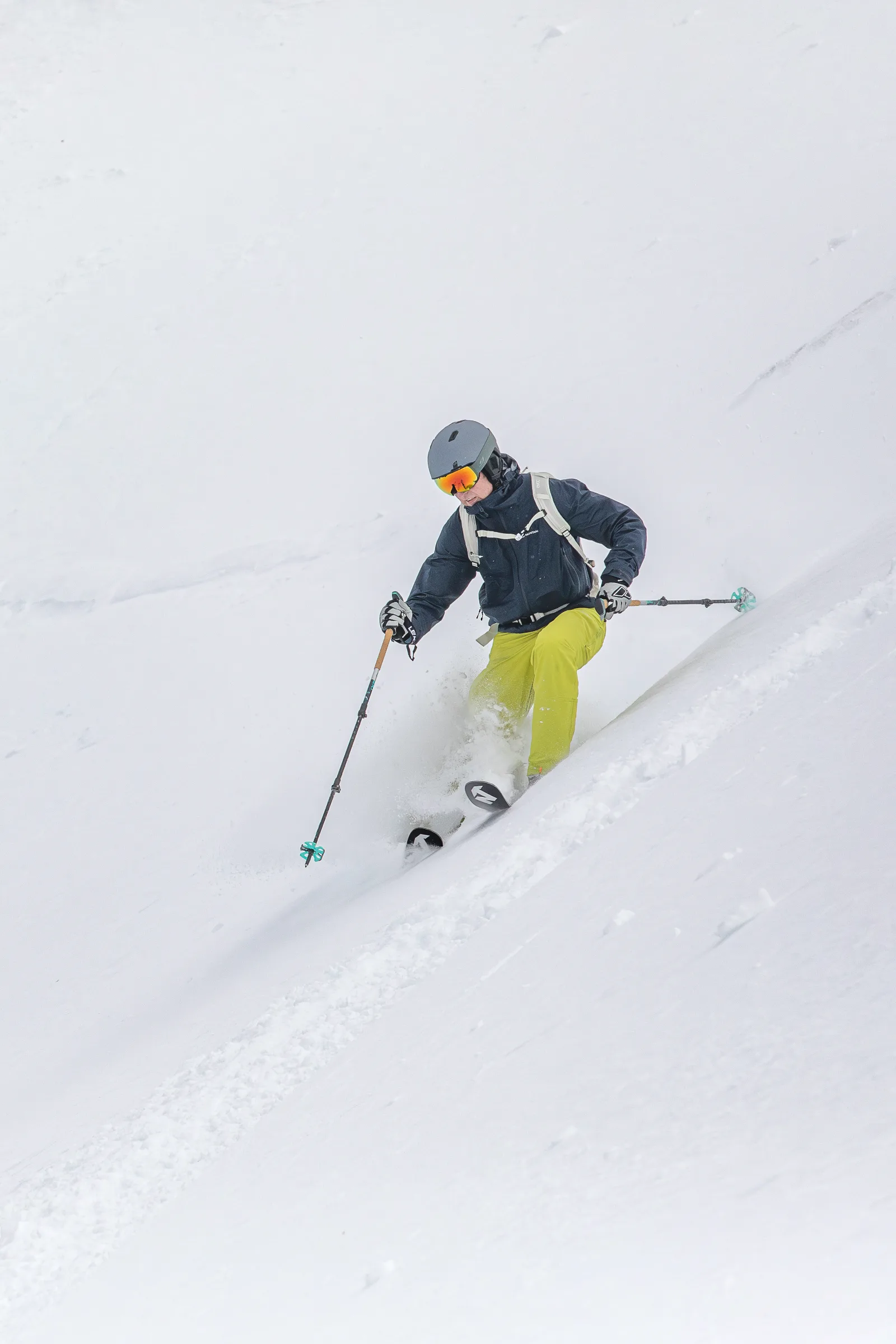 Christoph Öfele fährt mit gebeugten Knien durch tiefen Schnee am Hang, während Schnee um seine Ski und die gelbgrüne Hose aufwirbelt.