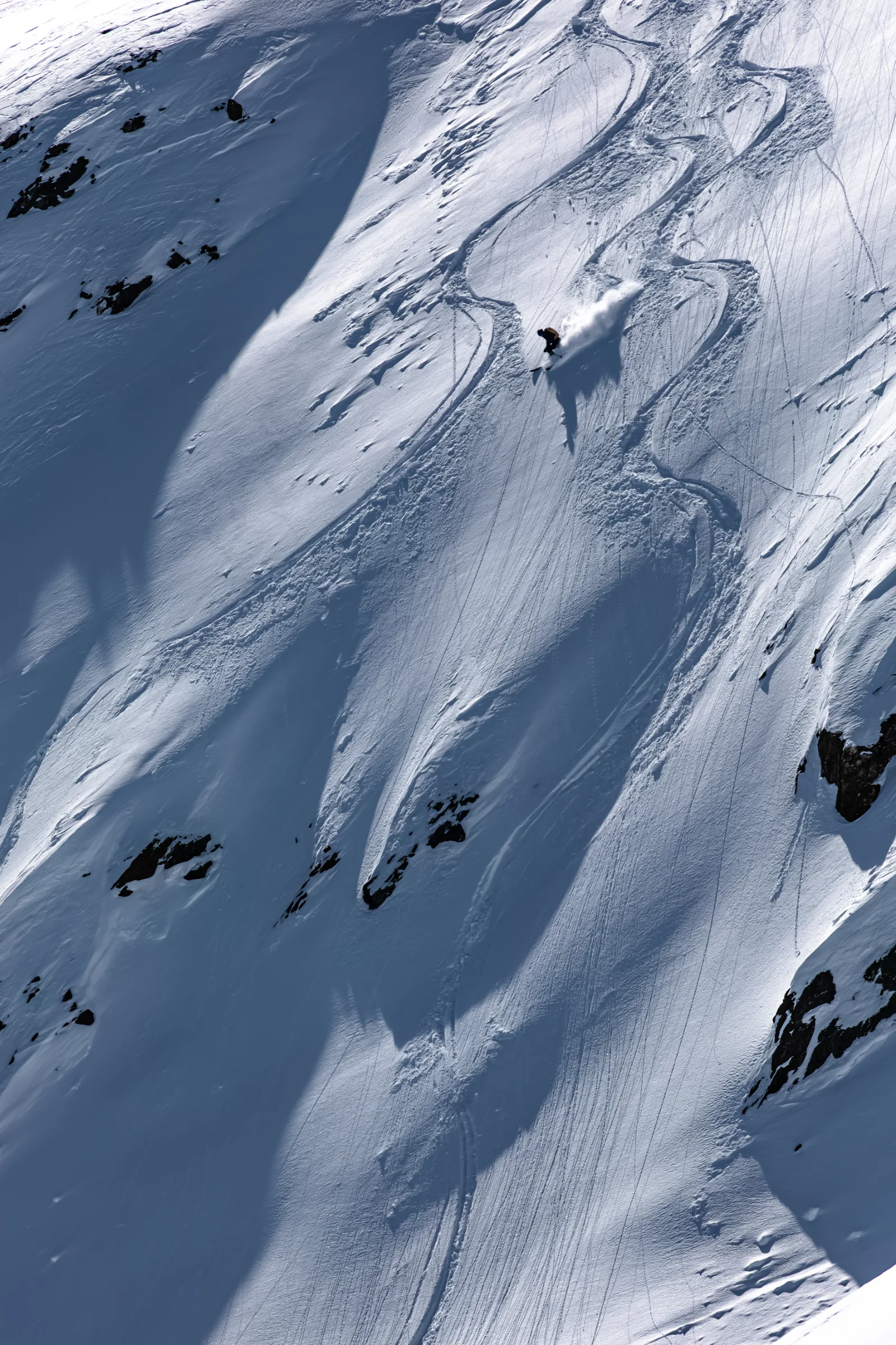 Christoph Öfele zieht eine Schneefahne über einen weiten, steilen Hang mit geschwungenen Skispuren, freiliegenden Felsen und langen Schatten.