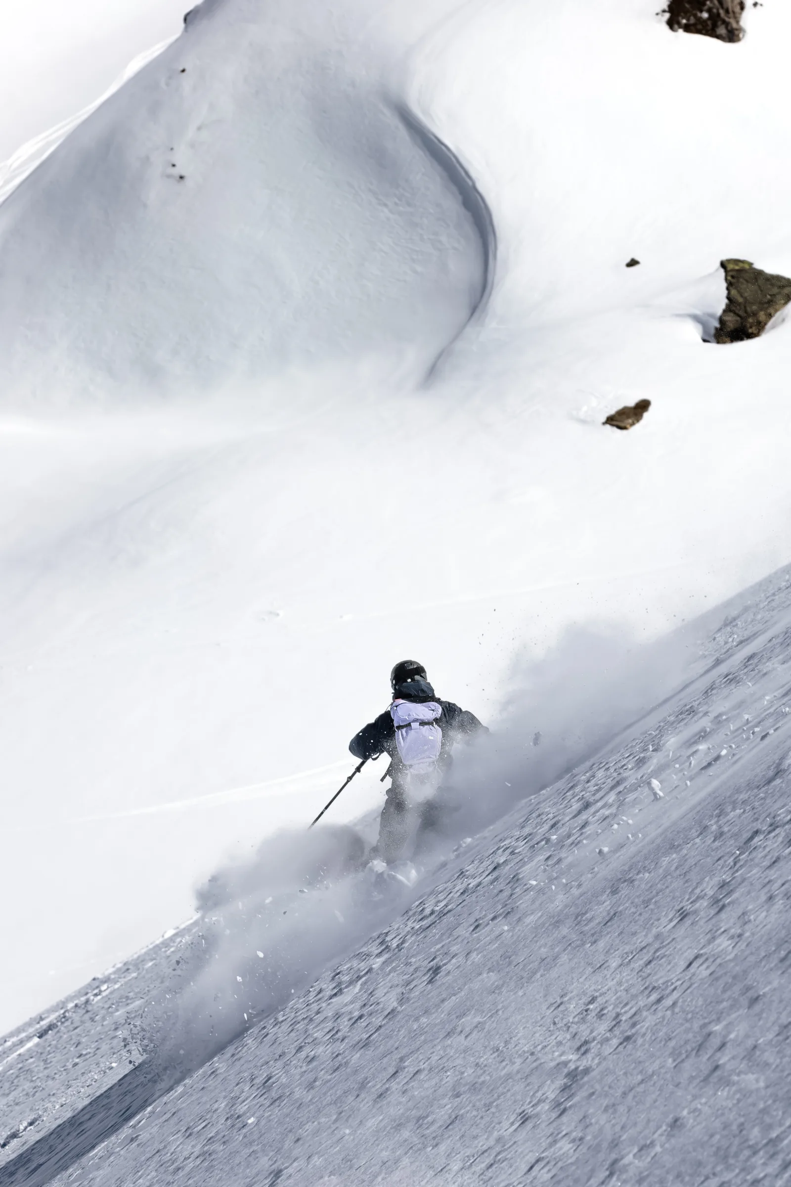 Von hinten gesehen fährt Christoph Öfele mit hellem Rucksack durch aufwirbelnden Schnee einen steilen Hang unterhalb geschwungener Schneeverwehungen hinab.
