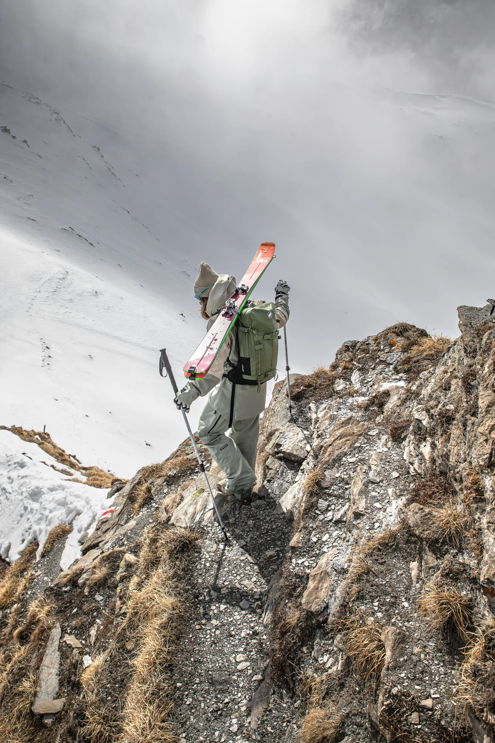 Claudia Kohler steigt mit Stöcken einen felsigen Pfad hinab, die Ski am grünen Rucksack befestigt, über einem wolkenverhangenen Schneehang.