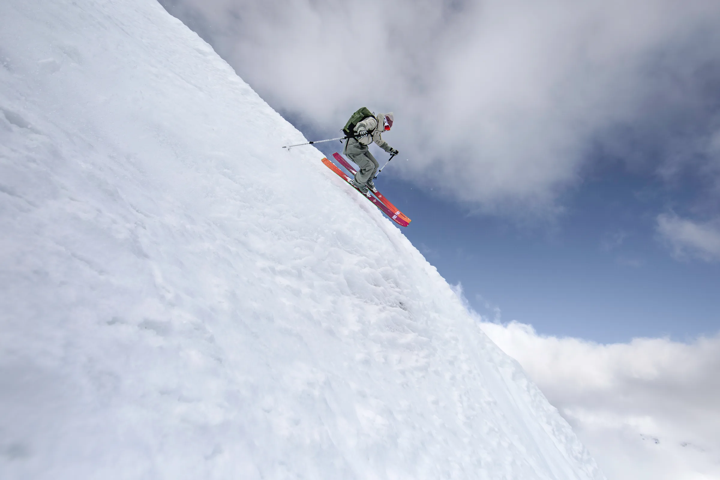 Claudia Kohler springt mit parallelen Ski über eine steile Schneekante vor blauem Himmel und weißen Wolken.