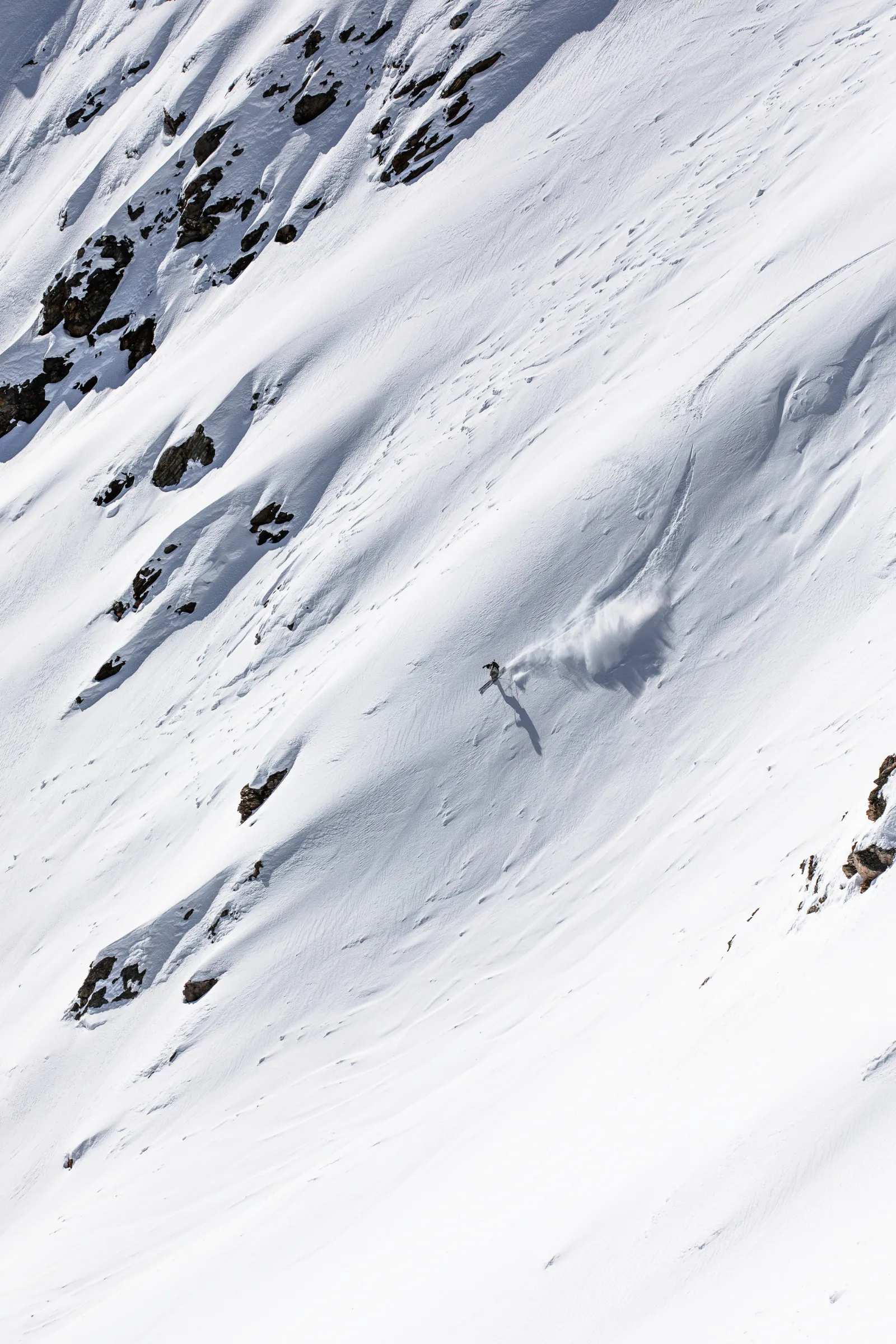 Claudia Kohler fährt zwischen freiliegenden Felsen einen weiten Schneehang hinab, mit einer großen Schneewolke und langem Schatten hinter sich.