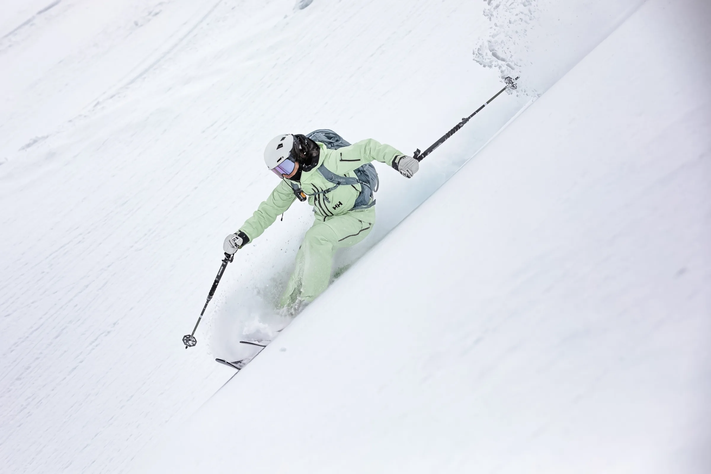 Claudia Kohler fährt in hellgrüner Skikleidung mit weißem Helm und grauem Rucksack durch tiefen Schnee an einem steilen Hang.