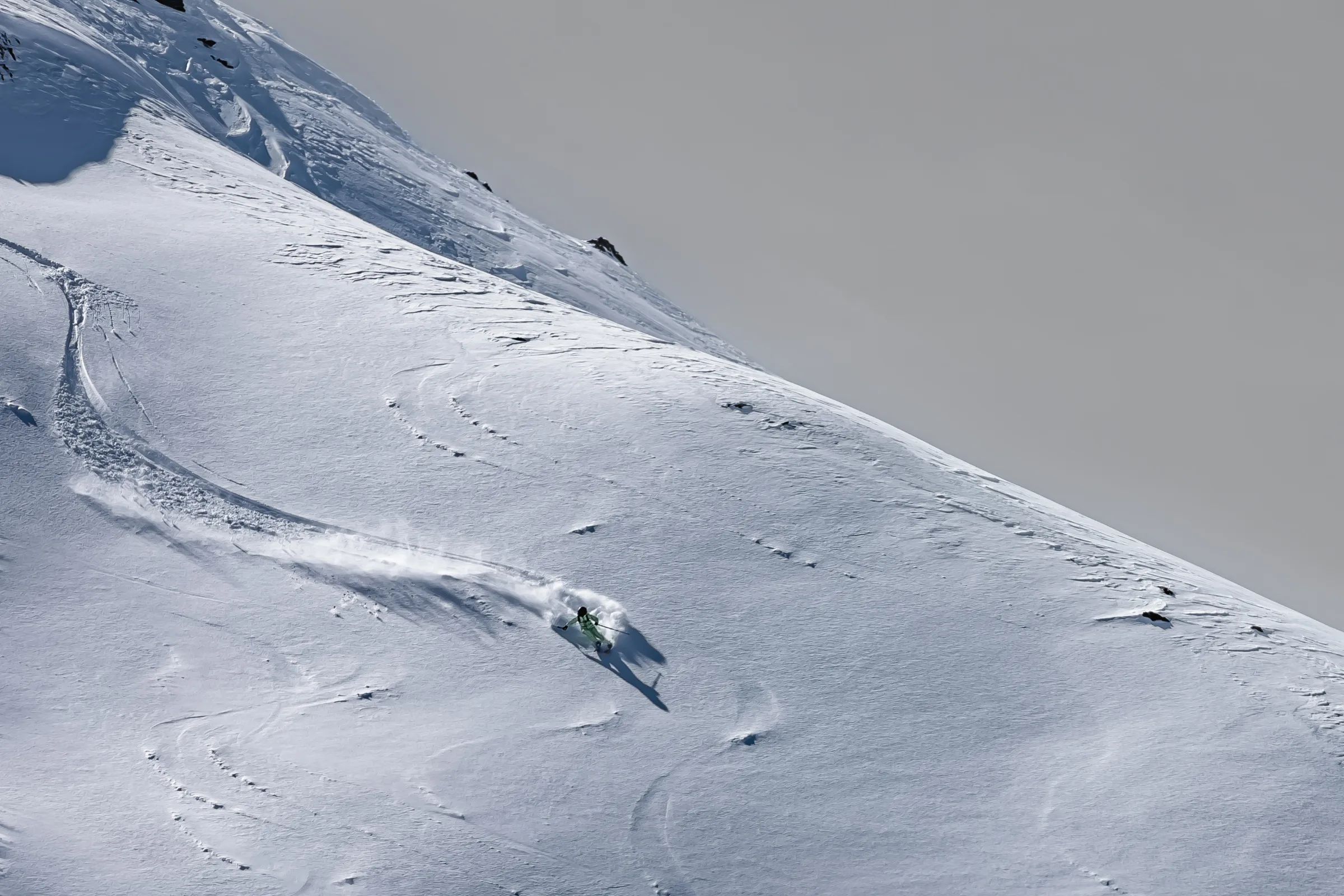 Claudia Kohler legt sich auf einem offenen Schneehang in die Kurve, während hinter ihr Schnee aufstäubt und ihr langer Schatten talwärts fällt.
