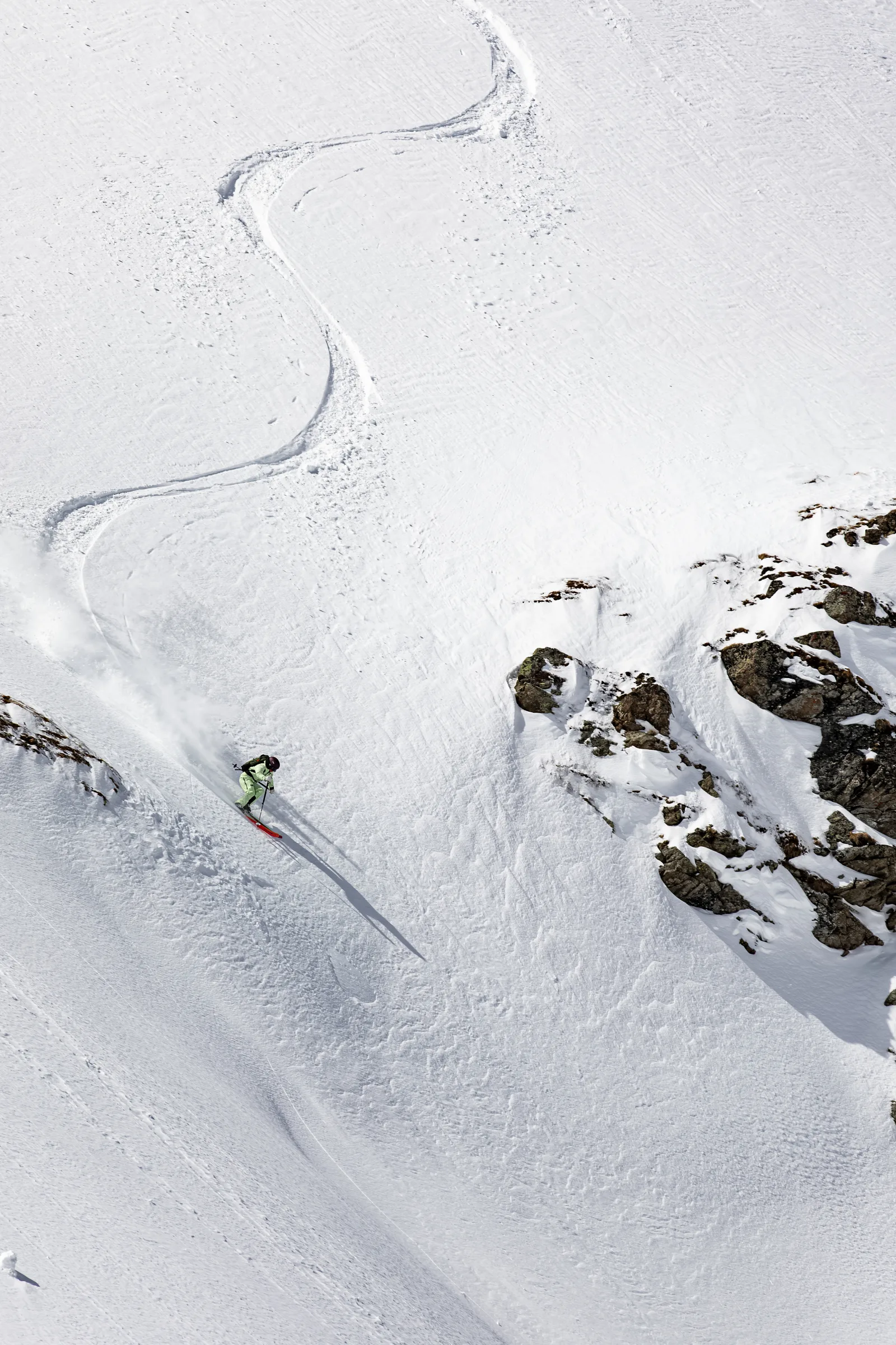 Claudia Kohler fährt neben Felsen einen sonnigen Schneehang hinab; oberhalb schlängelt sich ihre Skispur in mehreren Bögen über den Hang.