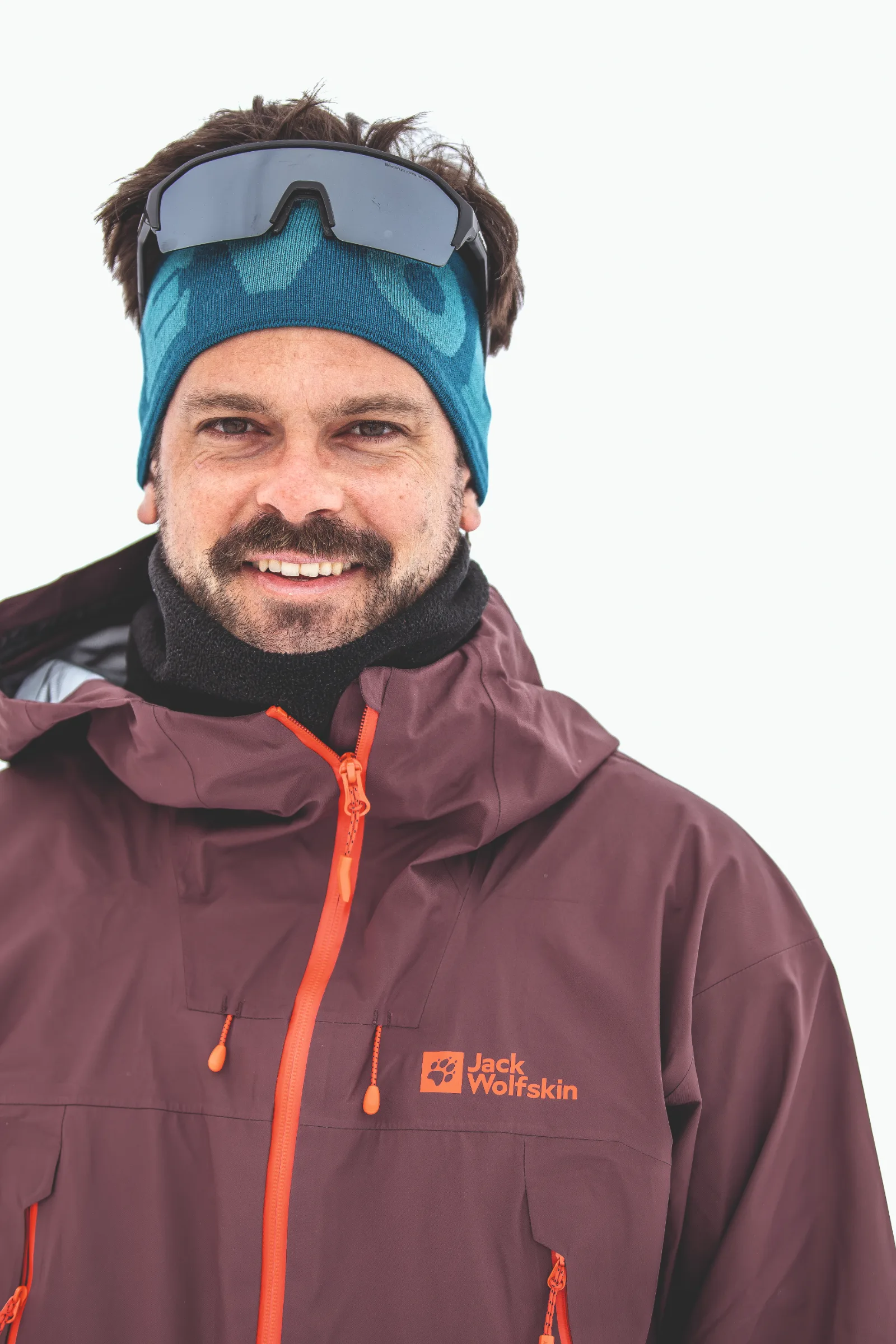 Florian Eigler lächelt vor weißem Hintergrund in die Kamera, mit blauem Stirnband, darauf sitzender Sonnenbrille und weinroter Jacke mit orangefarbenem Reißverschluss.