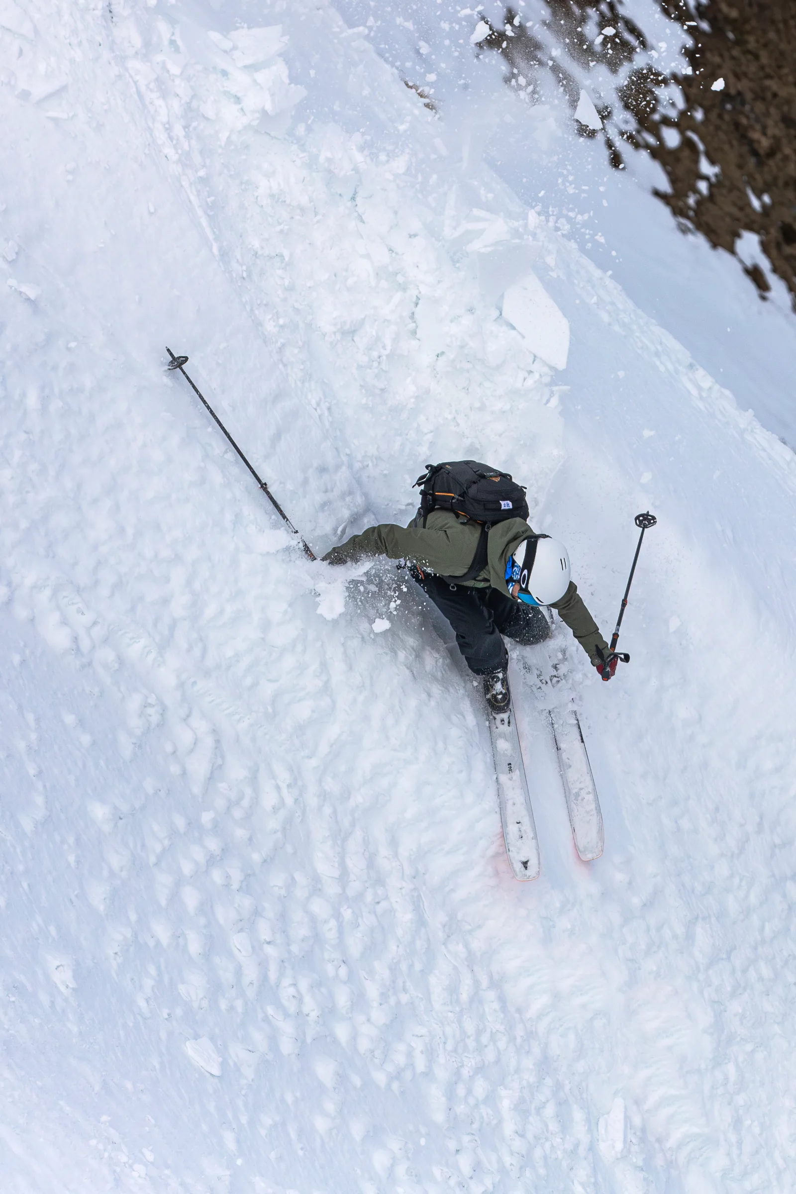 Von oben gesehen fährt Florian Eigler mit weißem Helm und Rucksack in tiefer Hocke durch aufgewirbelten Schnee neben einer Felswand.