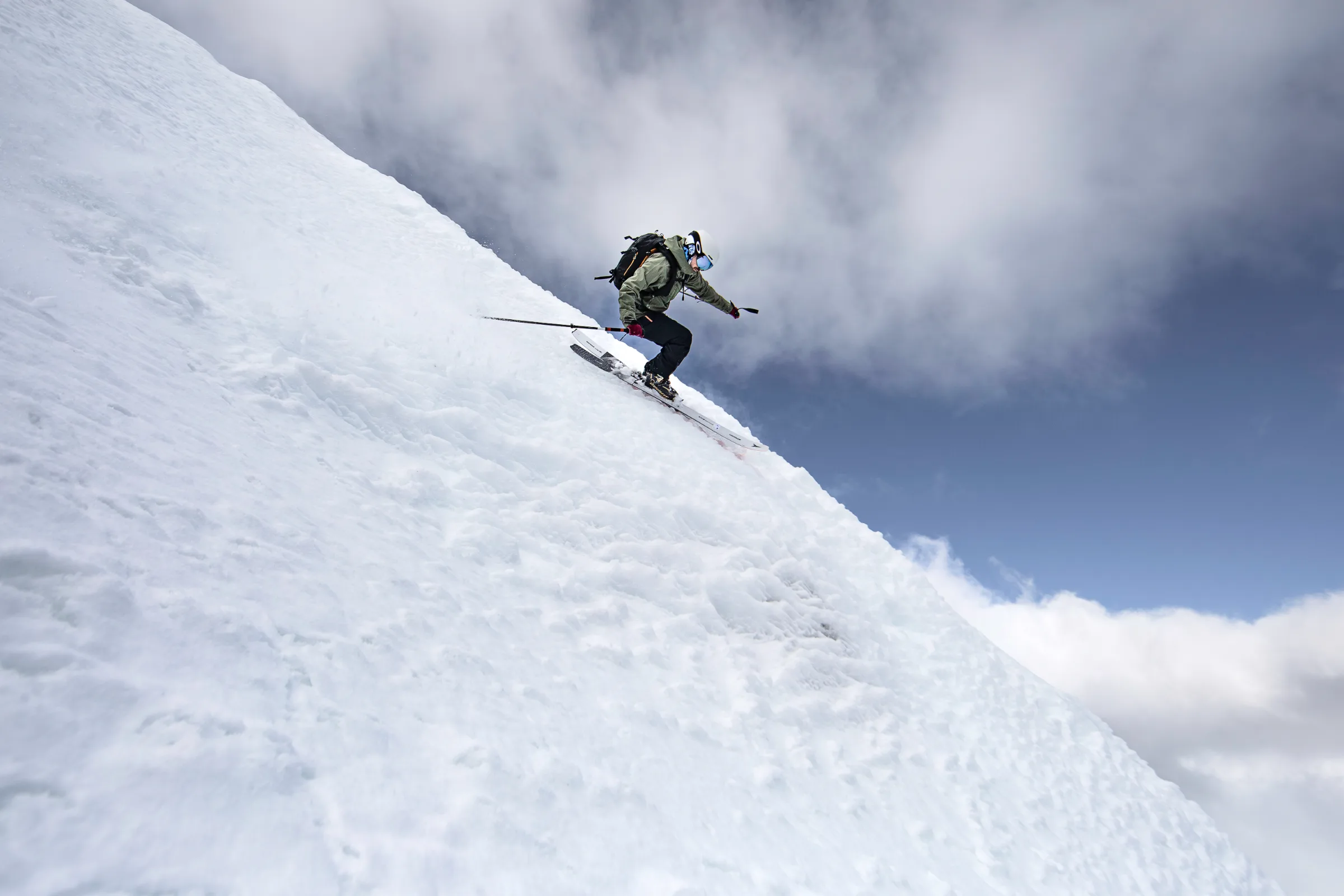 Florian Eigler fährt mit gebeugten Knien einen steilen Schneehang hinab, vor einem Himmel mit blauen Lücken und dichten Wolken.