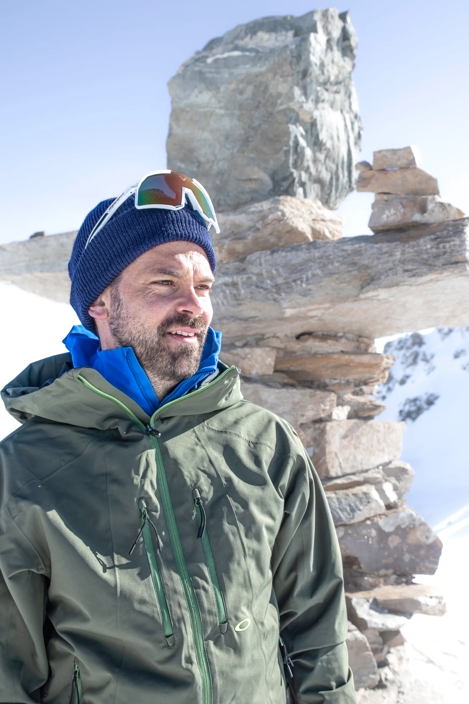 Florian Eigler blickt mit blauer Strickmütze und darauf sitzender Sonnenbrille lächelnd zur Seite, hinter ihm stehen aufgeschichtete Steine im Schnee.