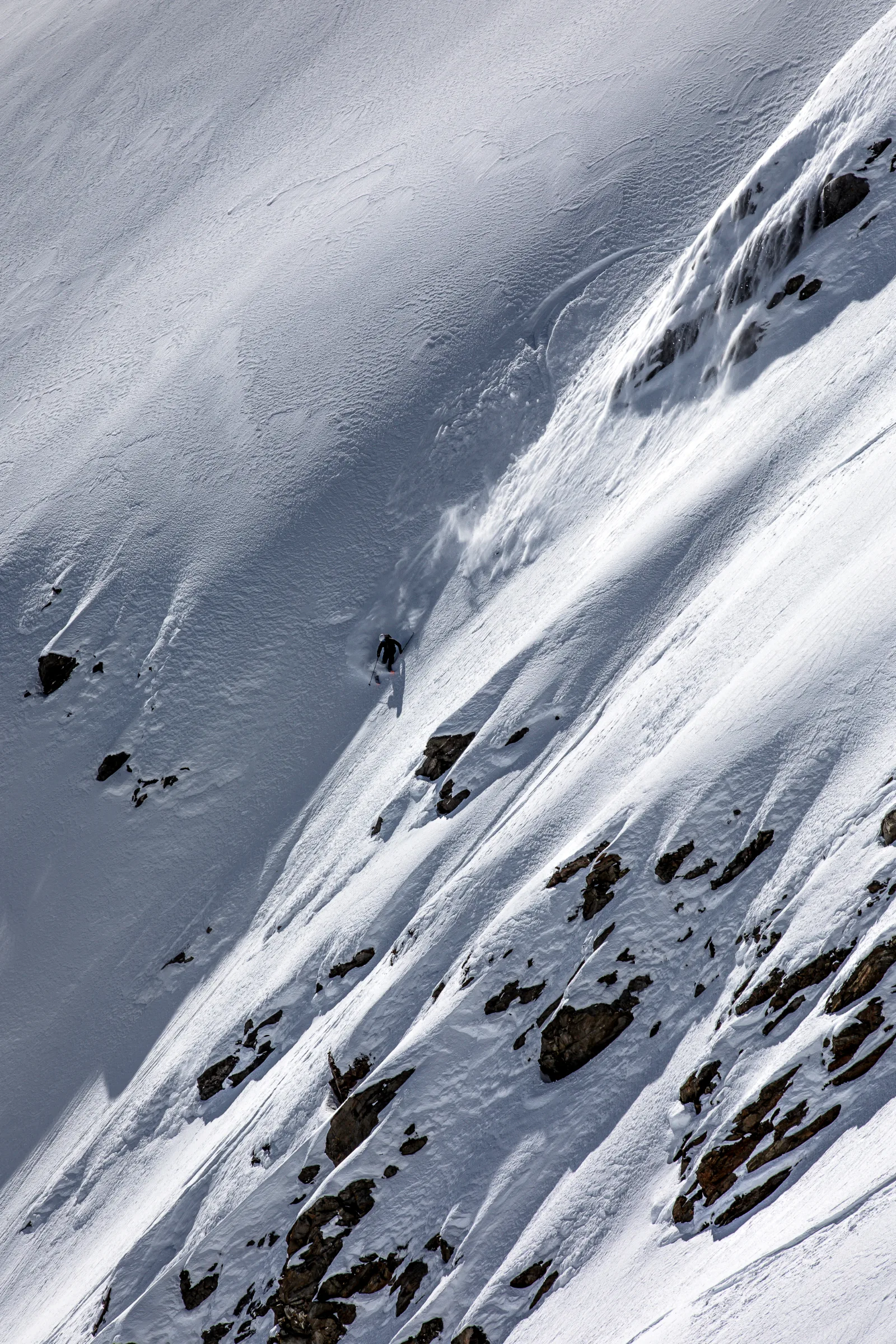 Florian Eigler fährt unterhalb einer gebogenen Skispur einen steilen Schneehang zwischen freiliegenden Felsen hinab, mit einer Schneefahne hinter sich.