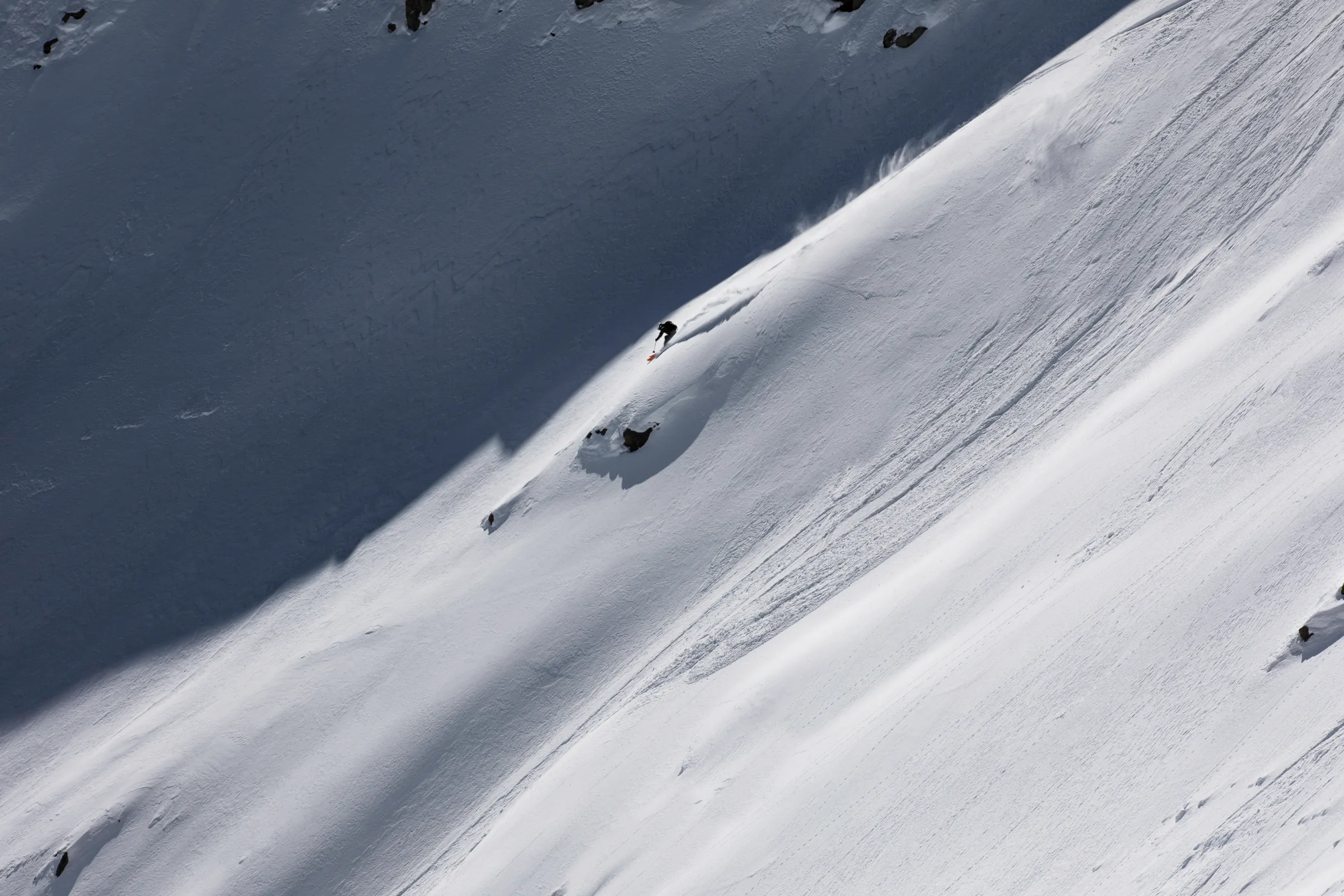 Florian Eigler fährt über einen weiten Schneehang entlang der Grenze zwischen Sonne und Schatten und hinterlässt eine Spur oberhalb einzelner Felsen.