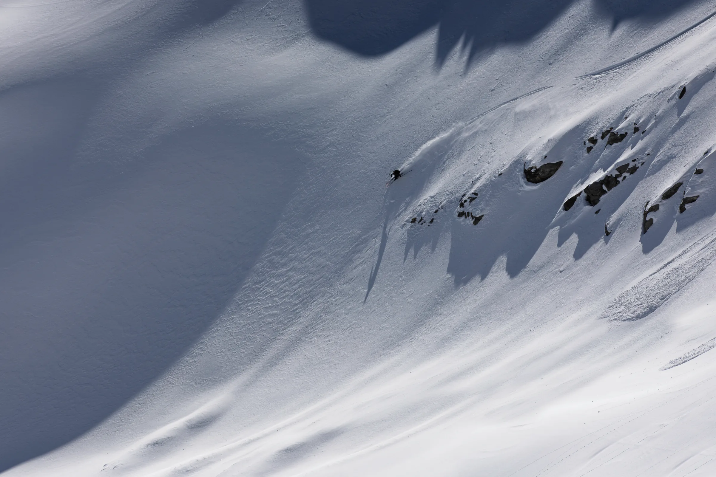 Florian Eigler zieht einen Schwung links einer Felsgruppe, deren lange Schatten über den Schneehang reichen, und wirbelt Schnee hinter sich auf.