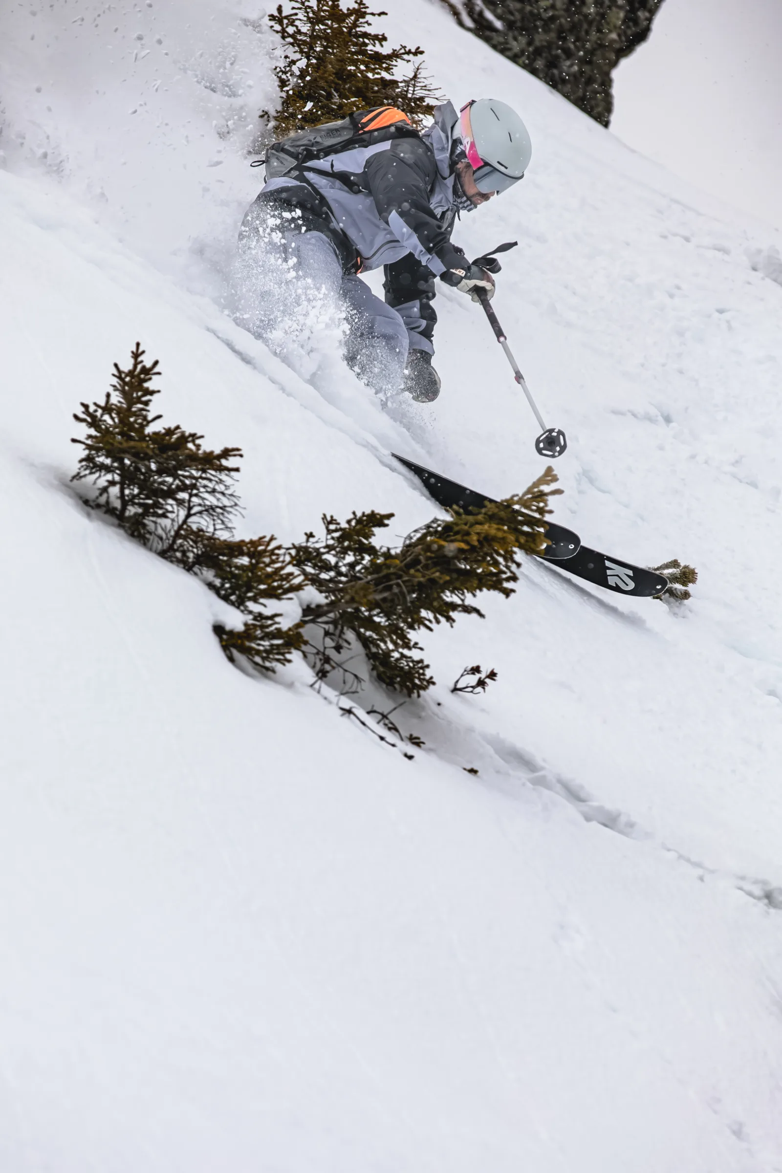 Florian Eigler fährt in gebeugter Haltung einen steilen Schneehang zwischen kleinen Nadelbäumen hinab und wirbelt Schnee auf.