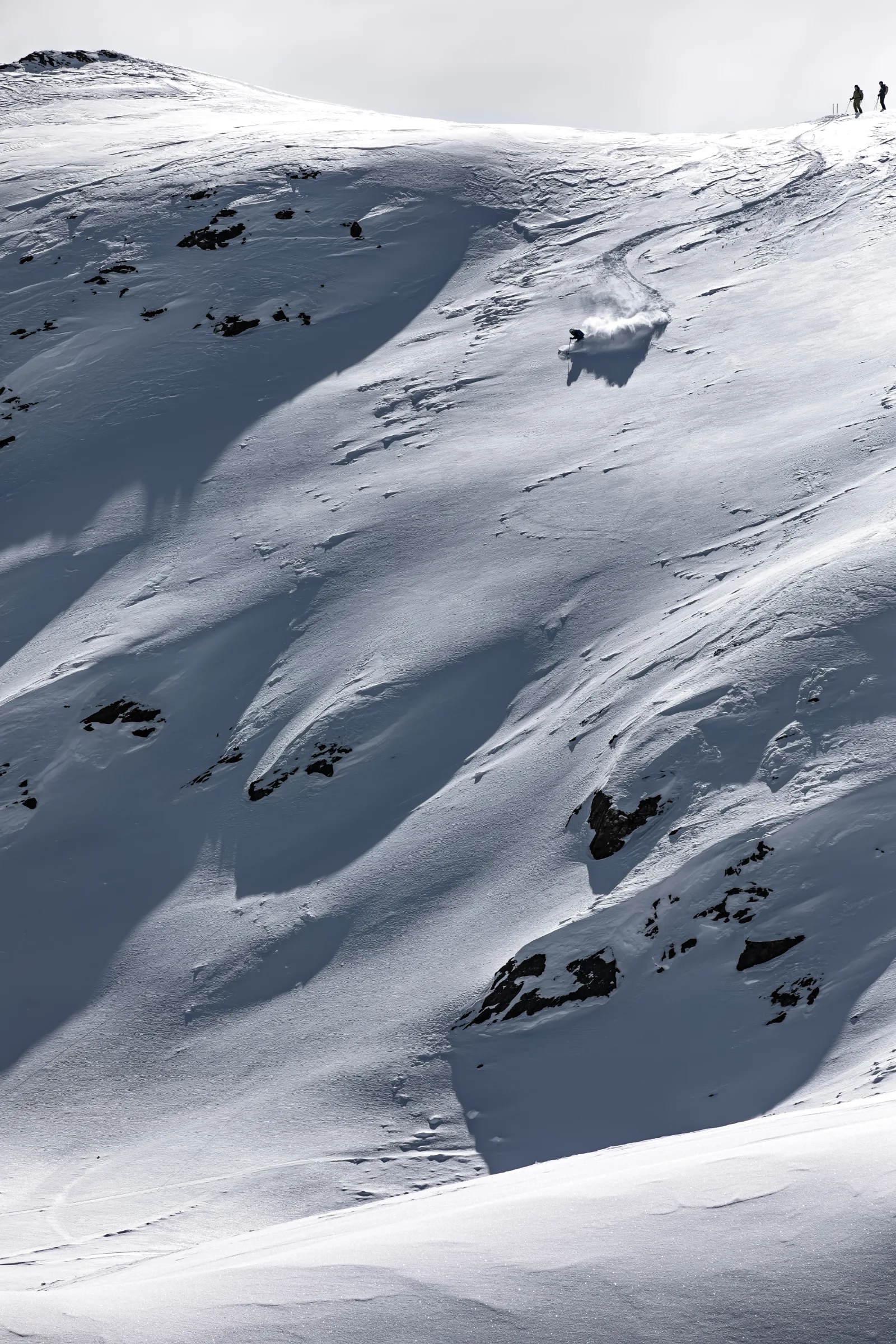 Florian Eigler zieht einen Schwung mit Schneefahne über einen weiten, felsdurchsetzten Hang in Lenzerheide, während zwei Personen oben am Grat stehen.