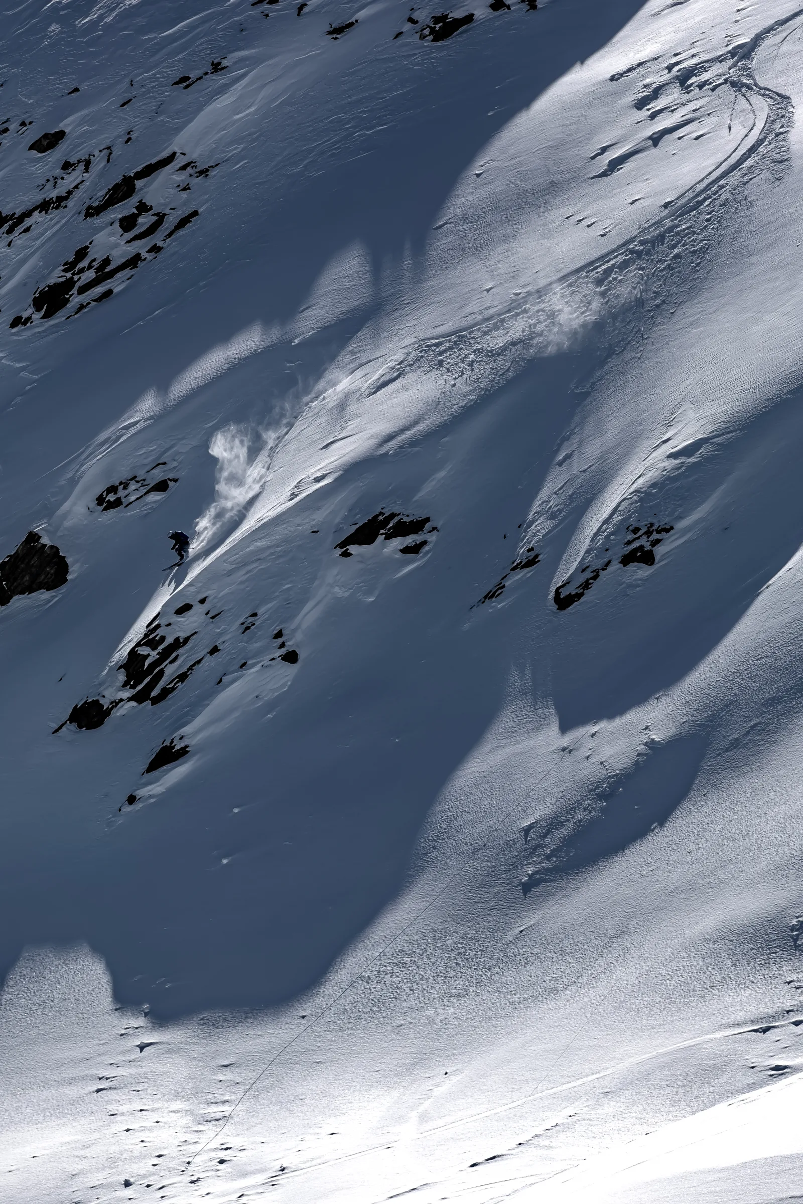 Florian Eigler fährt zwischen freiliegenden Felsen einen steilen, teils schattigen Schneehang hinab, hinter ihm eine lange Kurvenspur und aufgewirbelter Schnee.