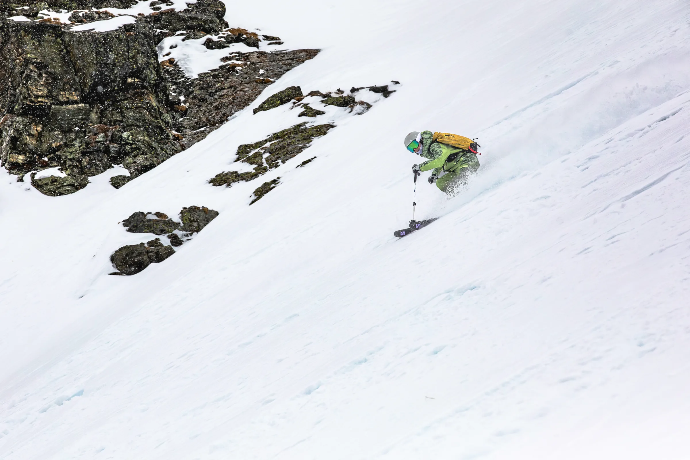 Martina Wengenmeir fährt neben einer dunklen Felswand über einen weiten, steilen Schneehang und wirbelt dabei Schnee auf.