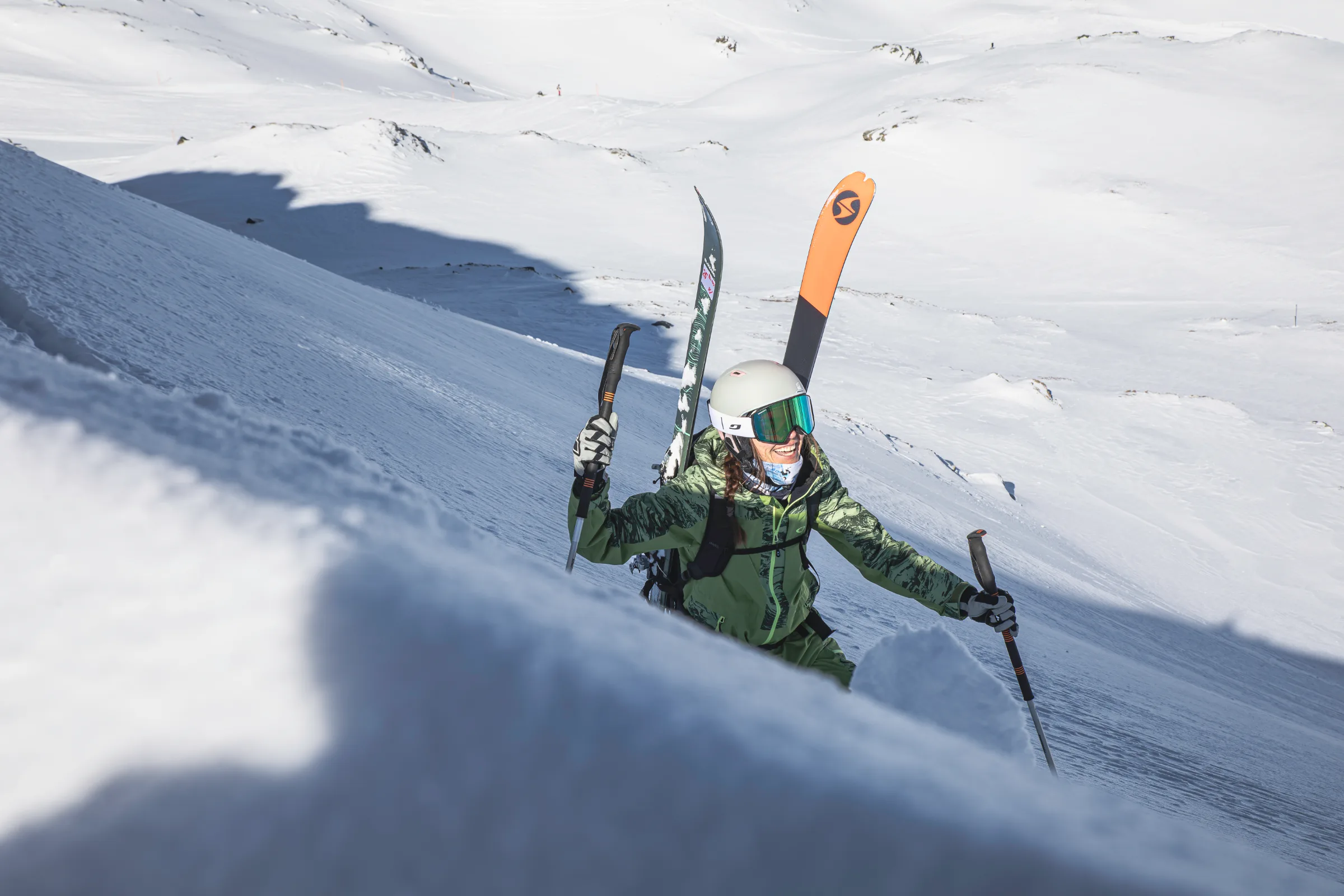 Martina Wengenmeir steigt mit Helm, Skibrille und Ski am Rucksack einen steilen Schneehang hinauf und stützt sich auf zwei Stöcke.