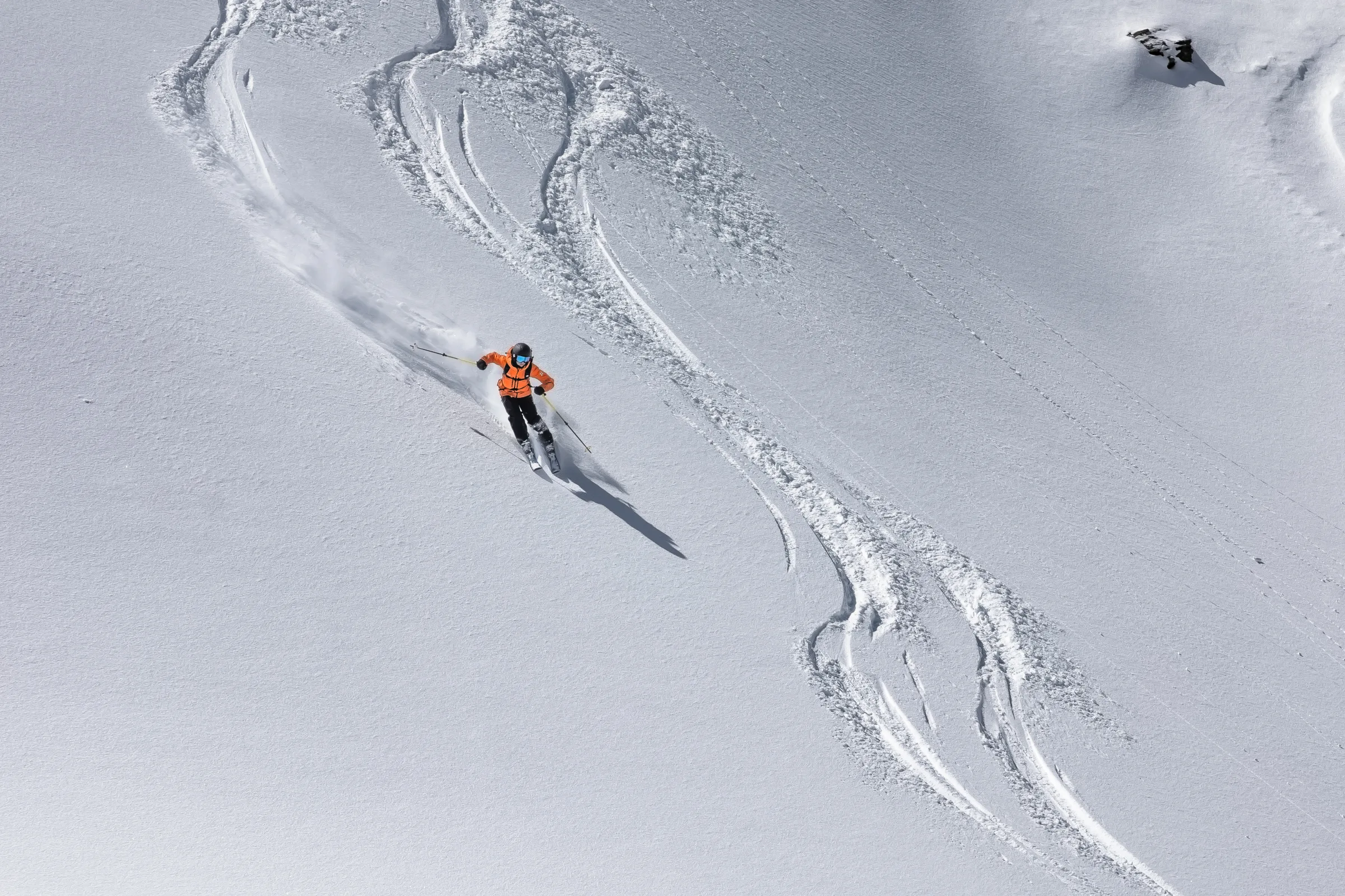 Martina Wengenmeir fährt in orangefarbener Jacke über einen offenen Schneehang, zieht eine Schneefahne hinter sich her und passiert geschwungene Skispuren.