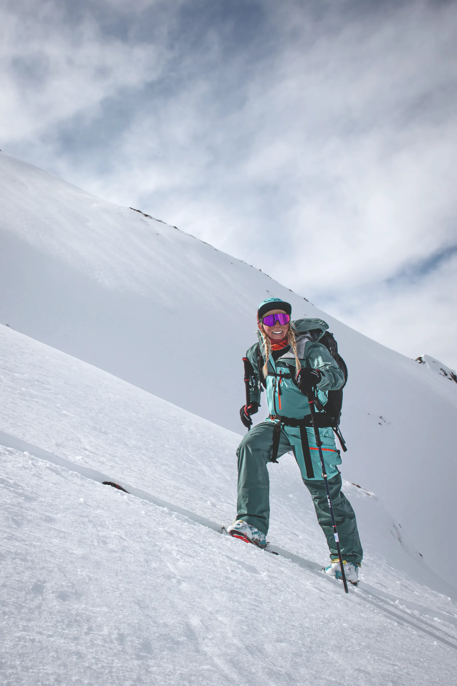 Michaela Casotti lächelt mit Ski, Stöcken und Rucksack an einem steilen Schneehang in Gastein, hinter ihr ein verschneiter Bergrücken.
