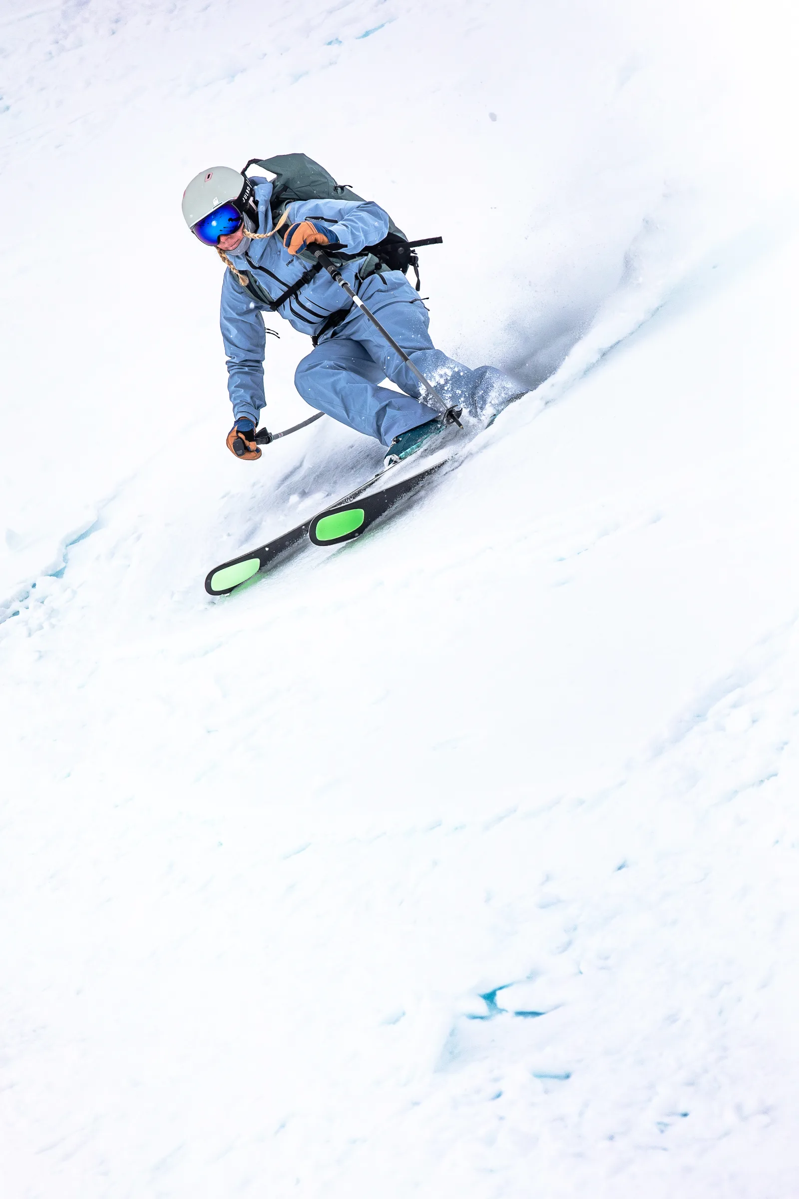 Michaela Casotti legt sich in hellblauer Skikleidung mit Helm und Rucksack in einen Schwung auf einem verschneiten Hang und wirbelt Schnee auf.