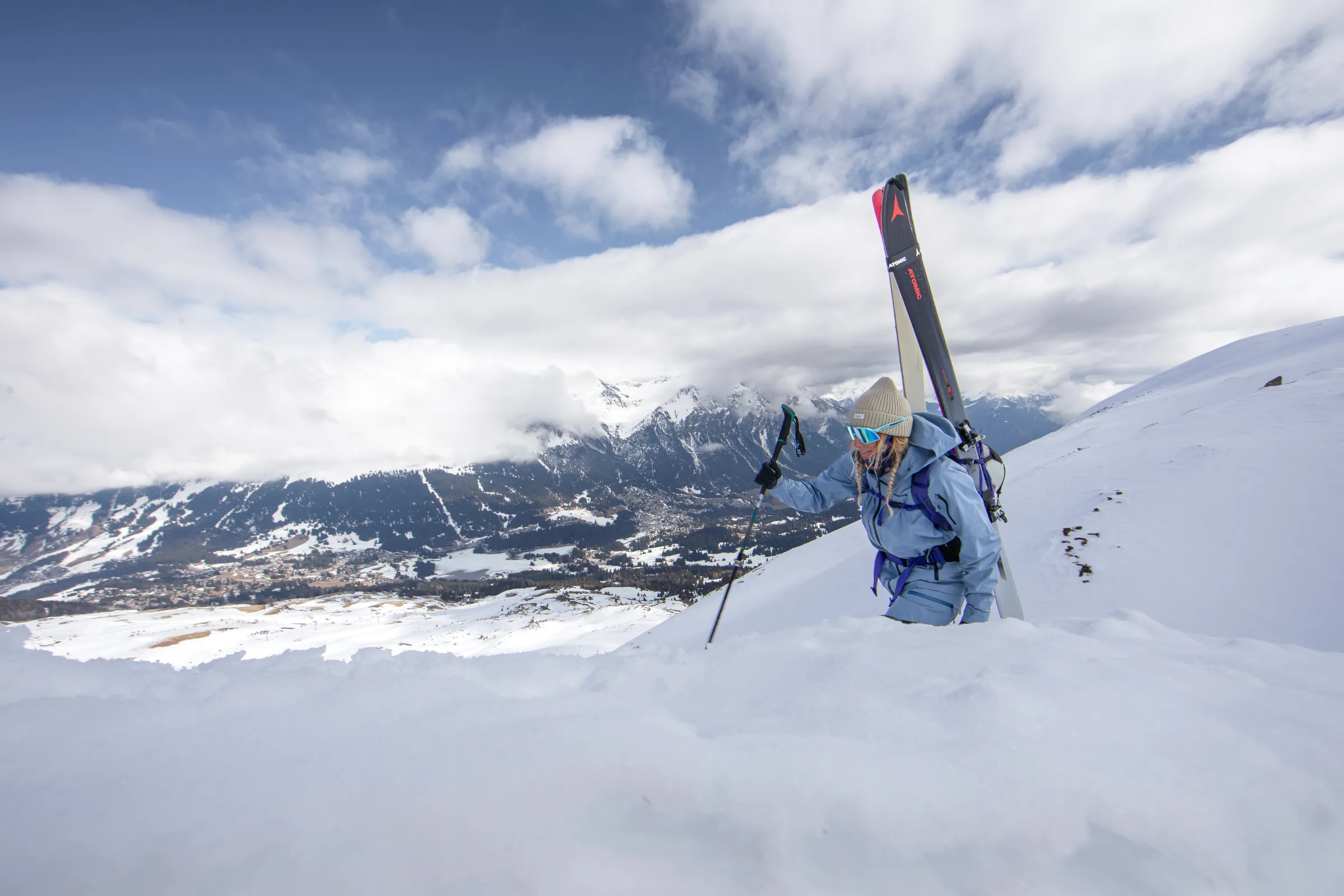 Michaela Casotti steigt mit Ski am Rucksack und einem Stock einen Schneehang über Lenzerheide hinauf, mit weitem Blick auf Tal und wolkenverhangene Berge.
