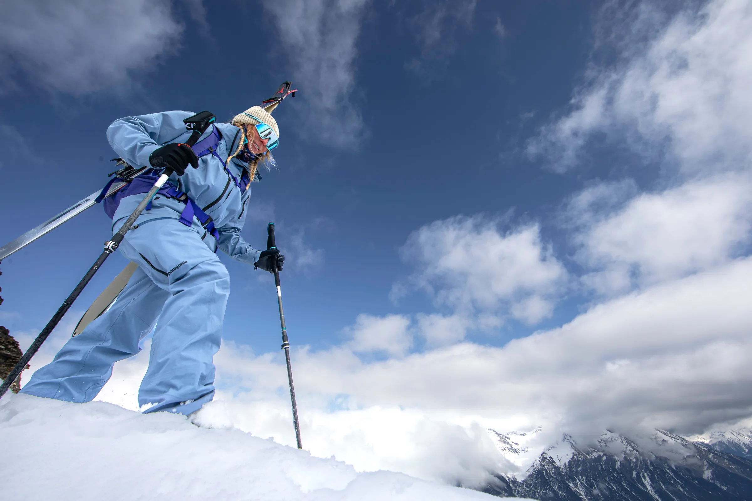 Aus tiefer Perspektive steigt Michaela Casotti mit Ski am Rucksack und zwei Stöcken über einen Schneehang vor blauem Himmel und weißen Wolken.