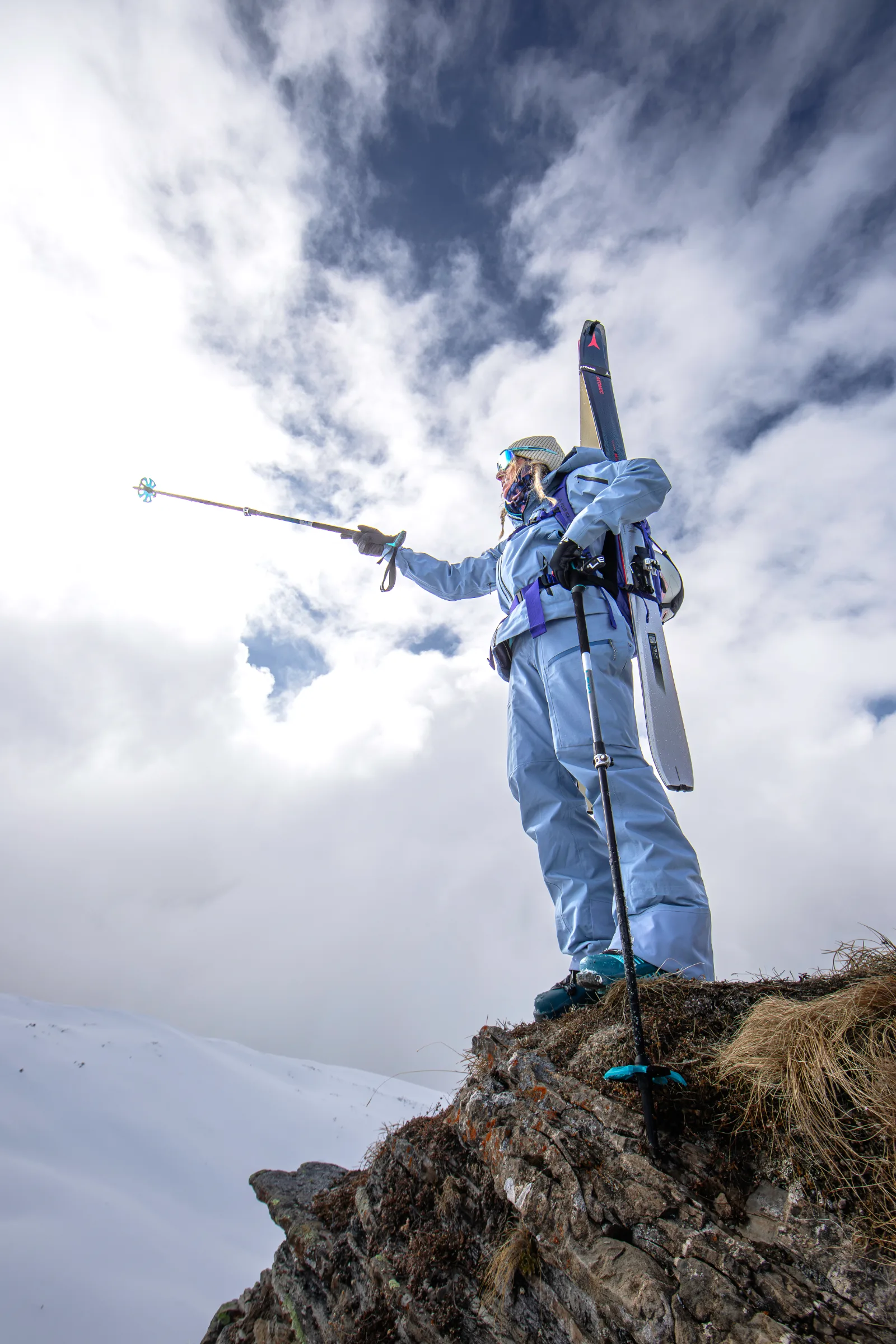 Michaela Casotti steht mit Ski am Rucksack auf einem grasigen Felsvorsprung und hält einen Stock nach links ausgestreckt vor einem wolkigen Himmel.