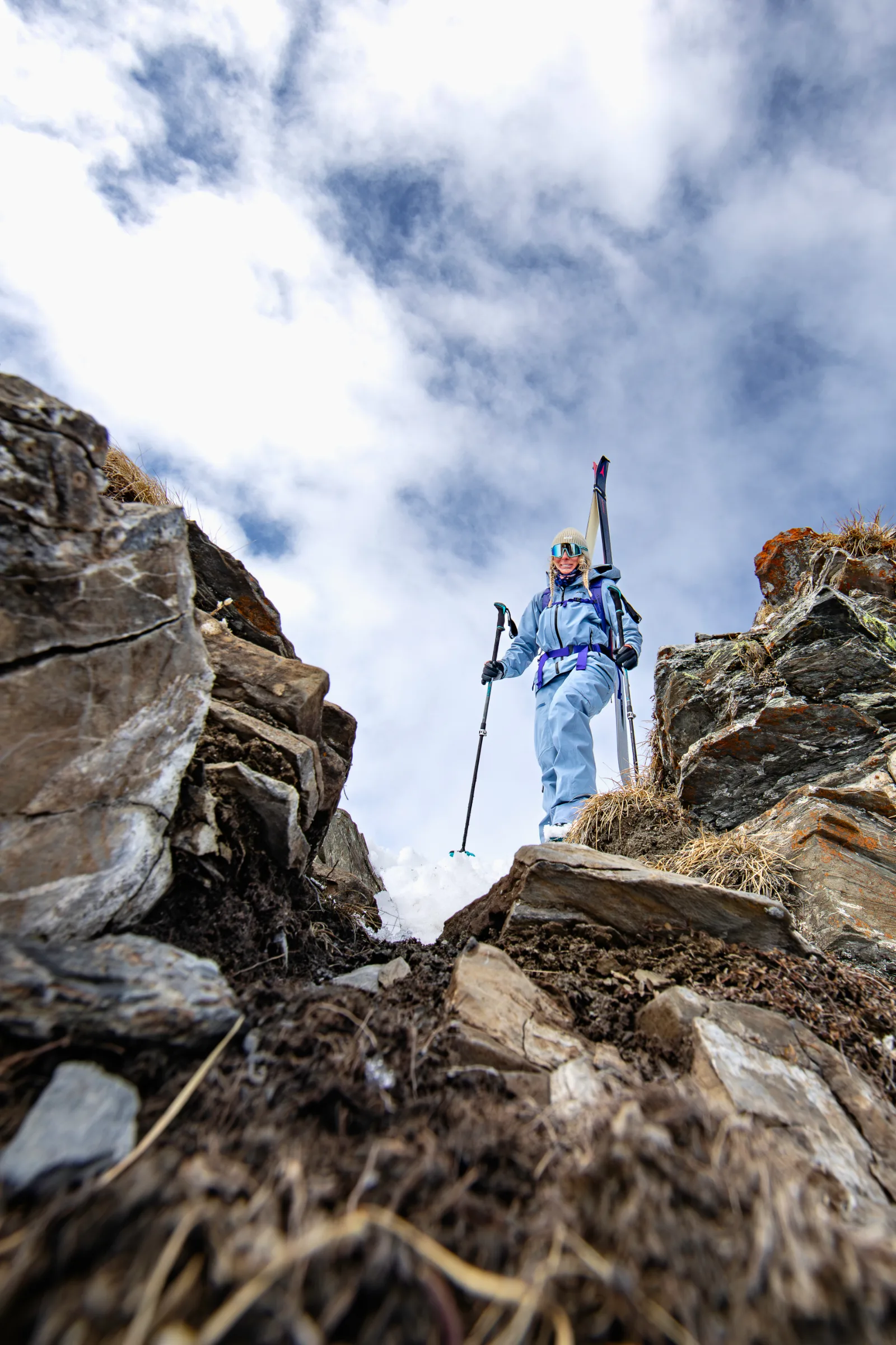 Michaela Casotti steht in hellblauer Skikleidung mit Ski am Rucksack und Stöcken zwischen schneebedeckten Felsen unter wolkigem Himmel.