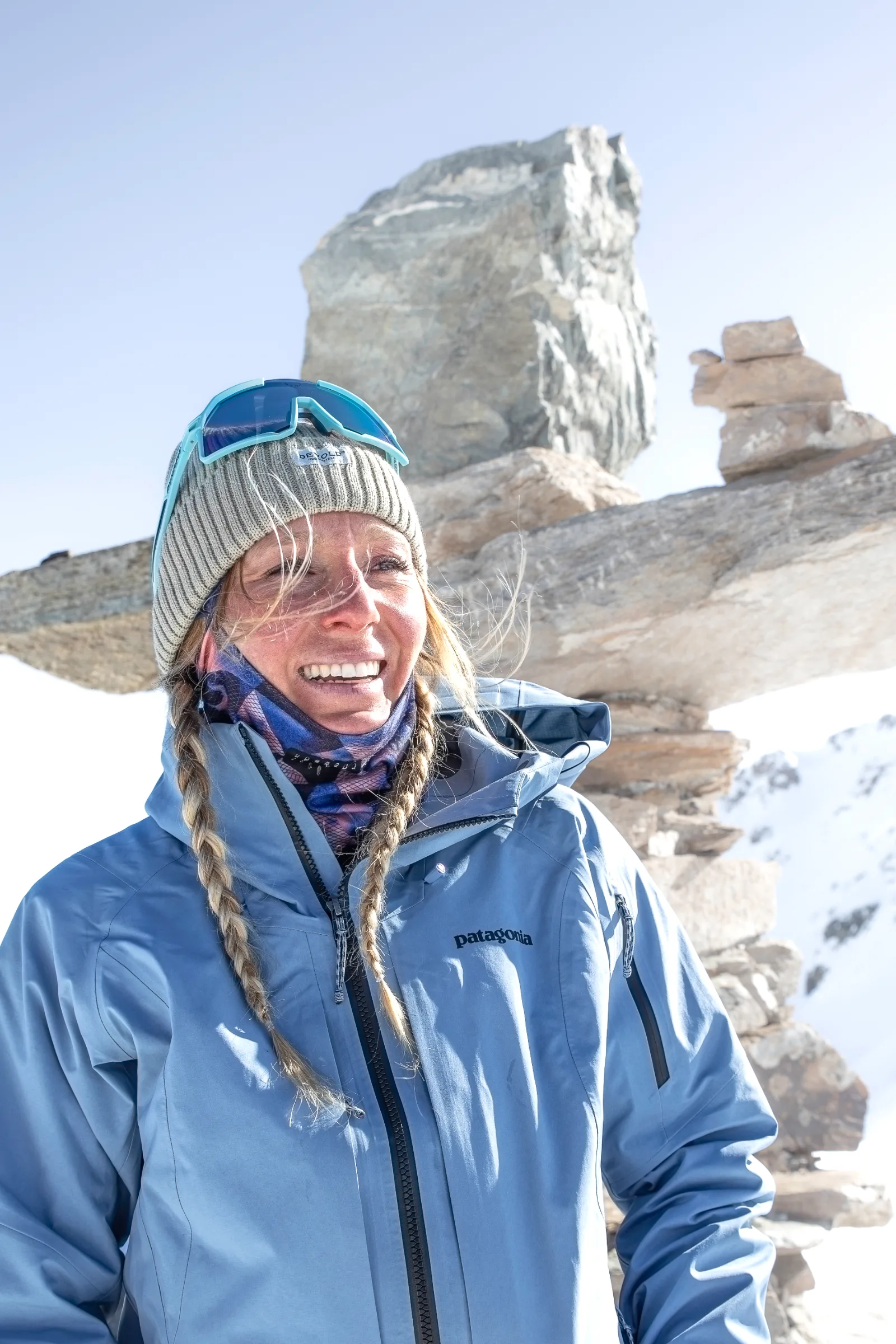 Michaela Casotti lächelt mit geflochtenen Zöpfen, Strickmütze und hochgeschobener Skibrille vor aufgeschichteten Steinplatten im Schnee.