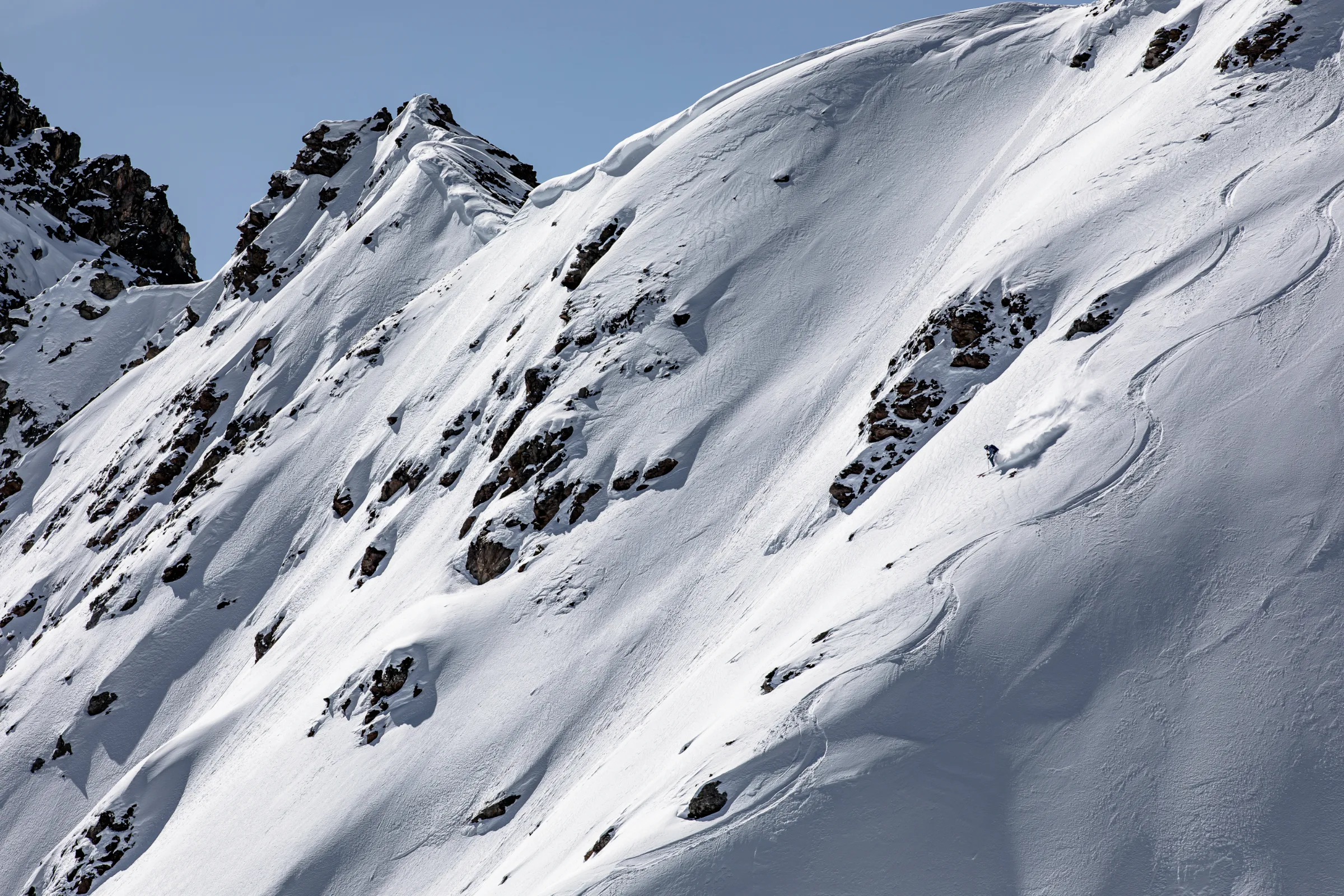 Michaela Casotti fährt unterhalb einer Felsgruppe über den steilen Schneehang und zieht eine Pulverschneewolke hinter sich her.