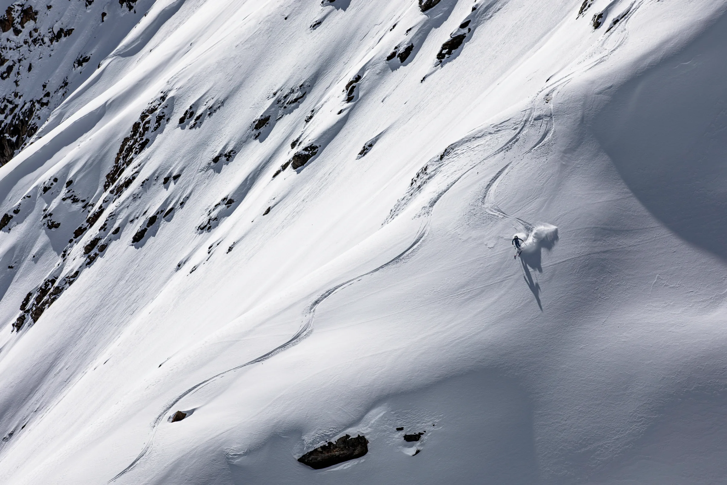 Michaela Casotti zieht einen Schwung durch einen offenen Schneehang; eine hohe Schneefahne und ihr langer Schatten begleiten sie.