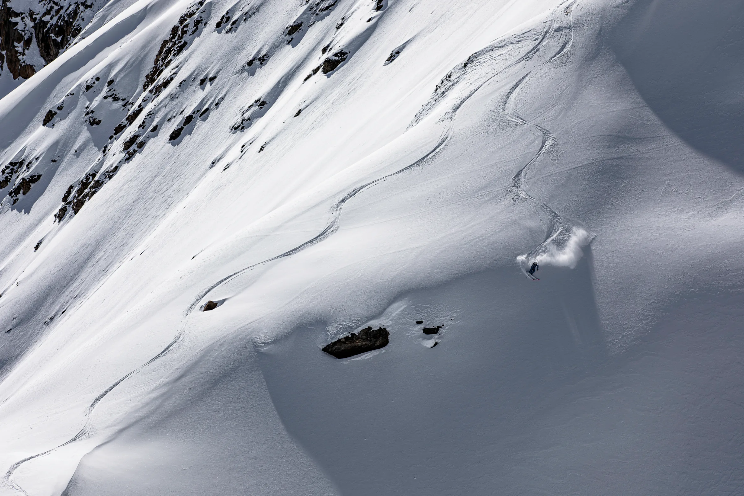 Michaela Casotti fährt einen weiten Schwung oberhalb freiliegender Felsen und hinterlässt eine geschwungene Spur mit aufspritzendem Pulverschnee.