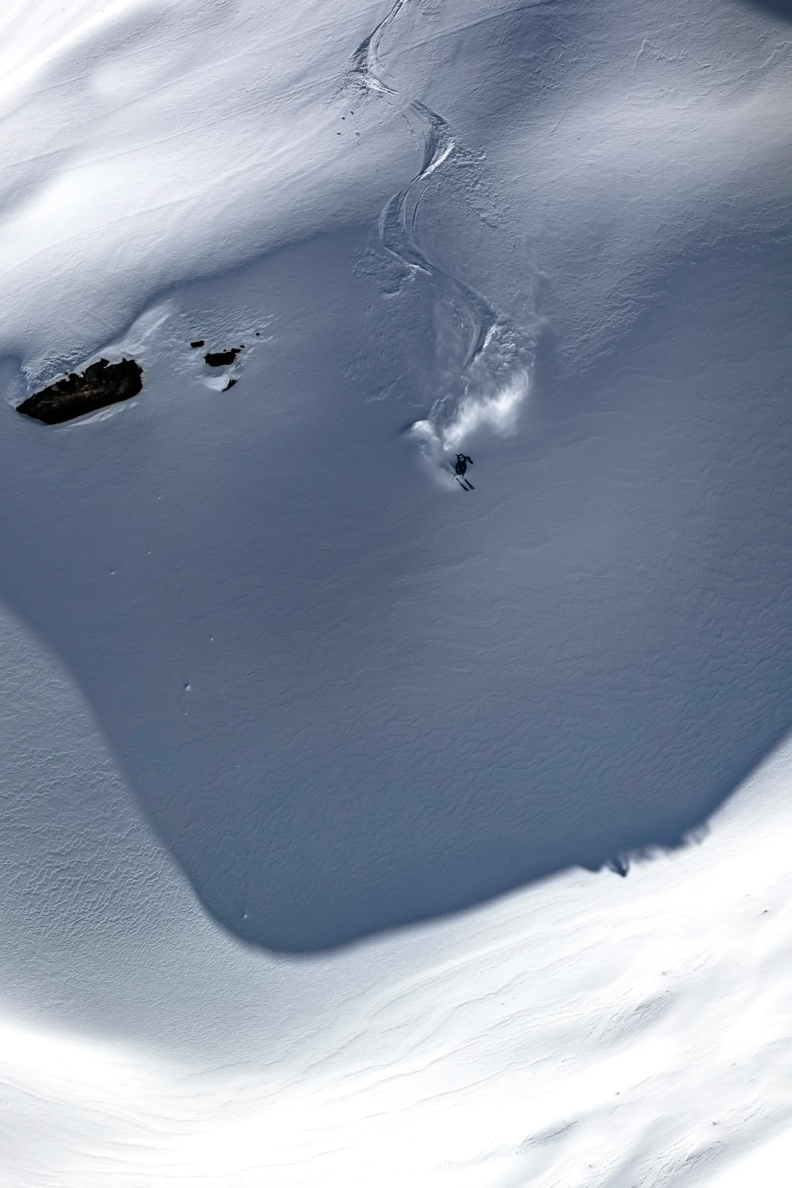 Michaela Casotti fährt unterhalb geschwungener Skispuren durch eine schattige Schneemulde, mit aufgewirbeltem Schnee hinter sich und Felsen links.